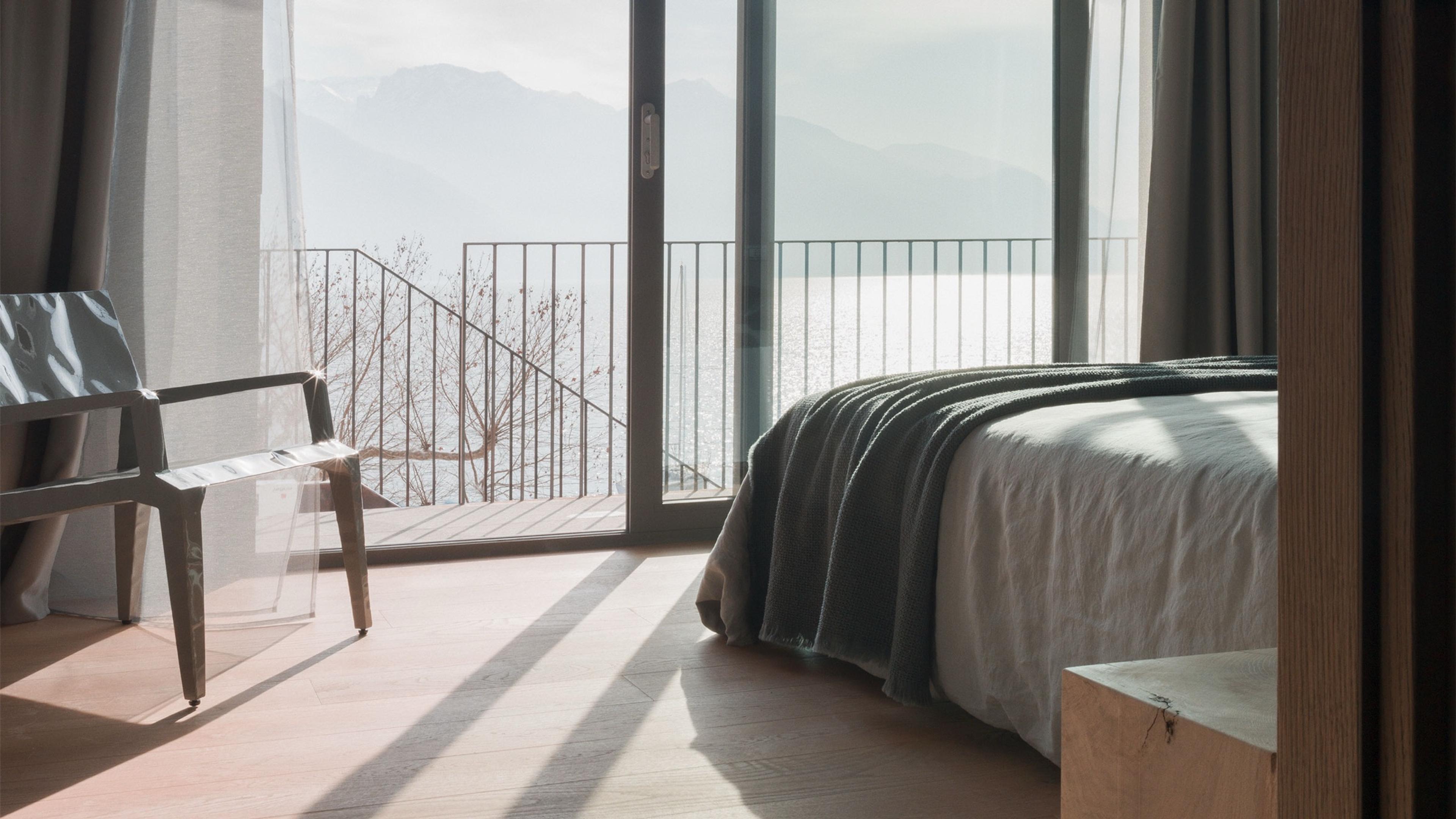 a bed with a grey throw faces open glass doors onto a balcony over the lake and mountains