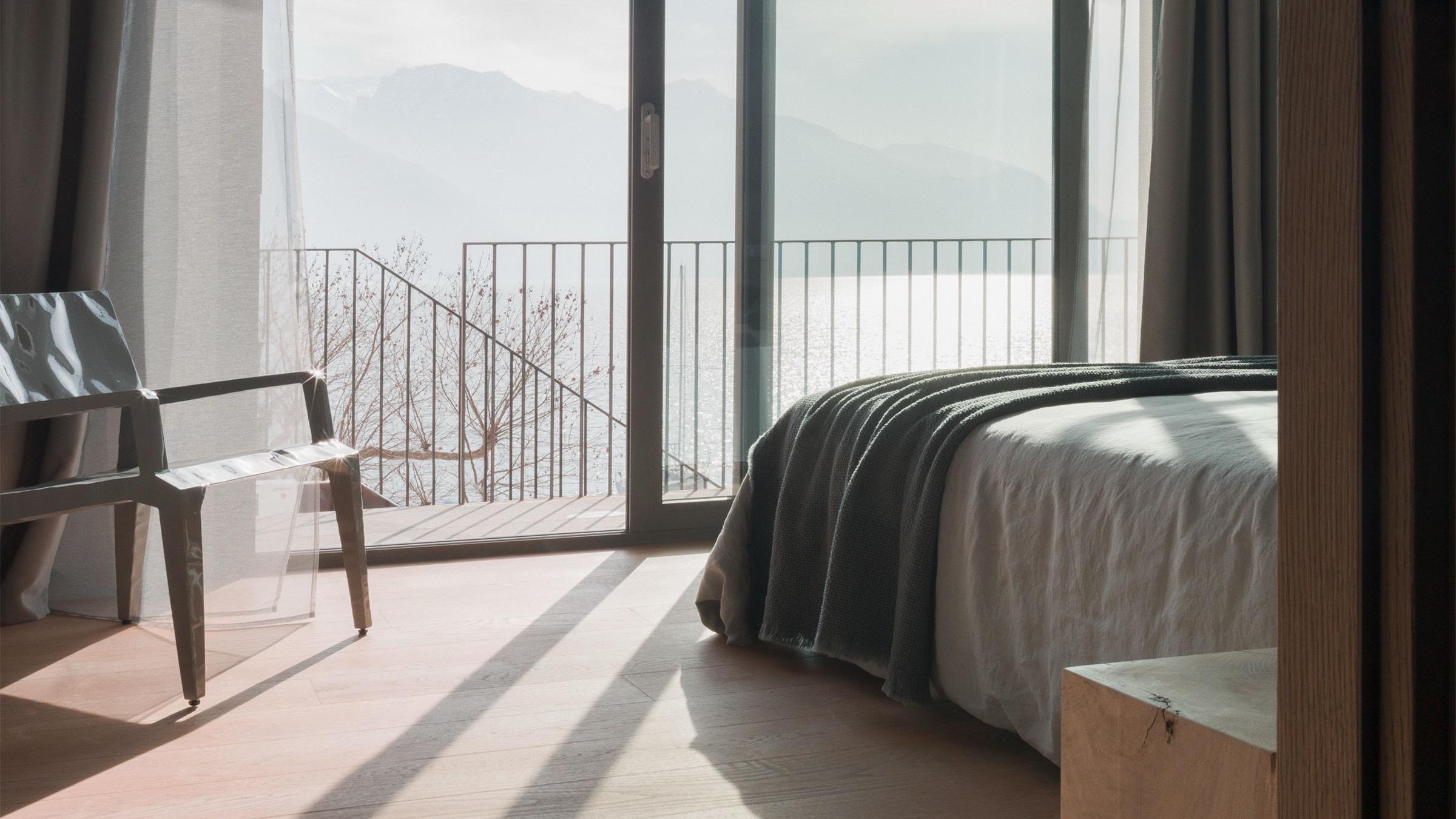 a bed with a grey throw faces open glass doors onto a balcony over the lake and mountains
