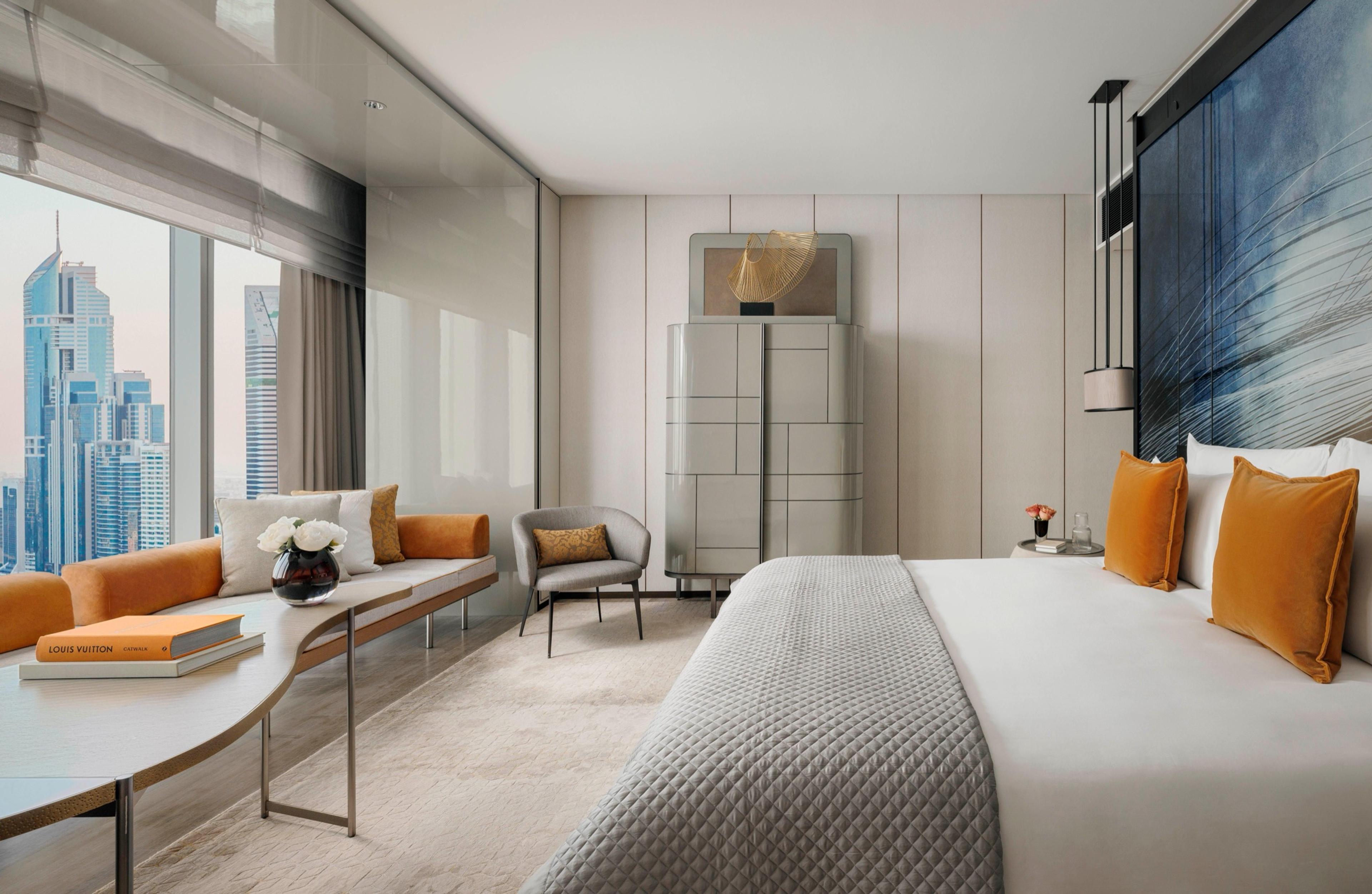 A bedroom with a grey upholstered bed topped in orange cushions, a dark patterned headboard panel, a round cream side table and a city view through the window.