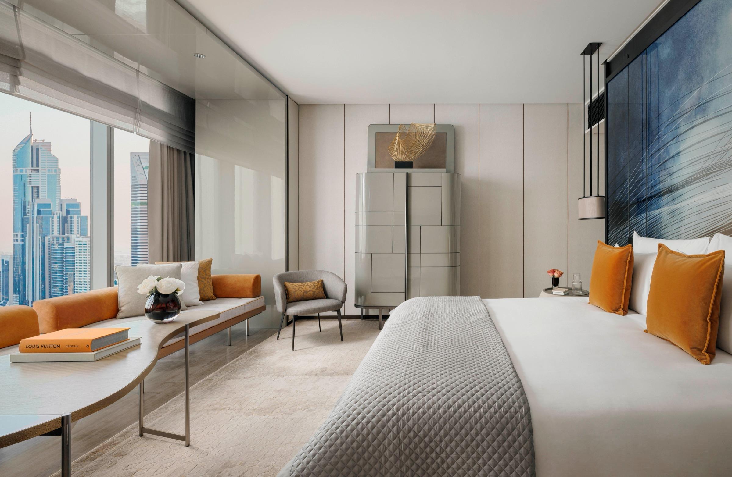 A bedroom with a grey upholstered bed topped in orange cushions, a dark patterned headboard panel, a round cream side table and a city view through the window.