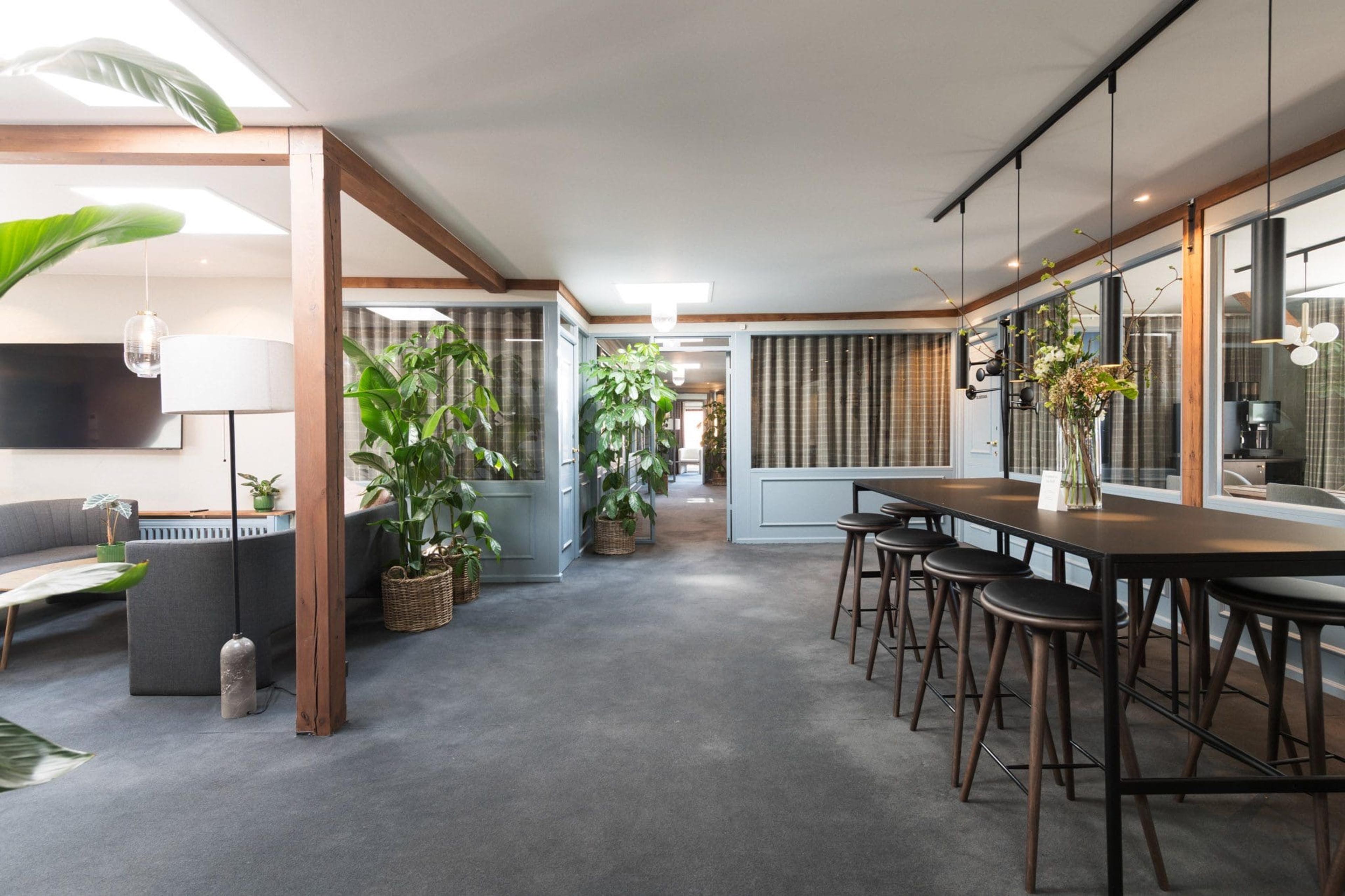 A lounge area with grey sofas, potted plants, a wooden-beamed ceiling and a long bar table lined with stools.
