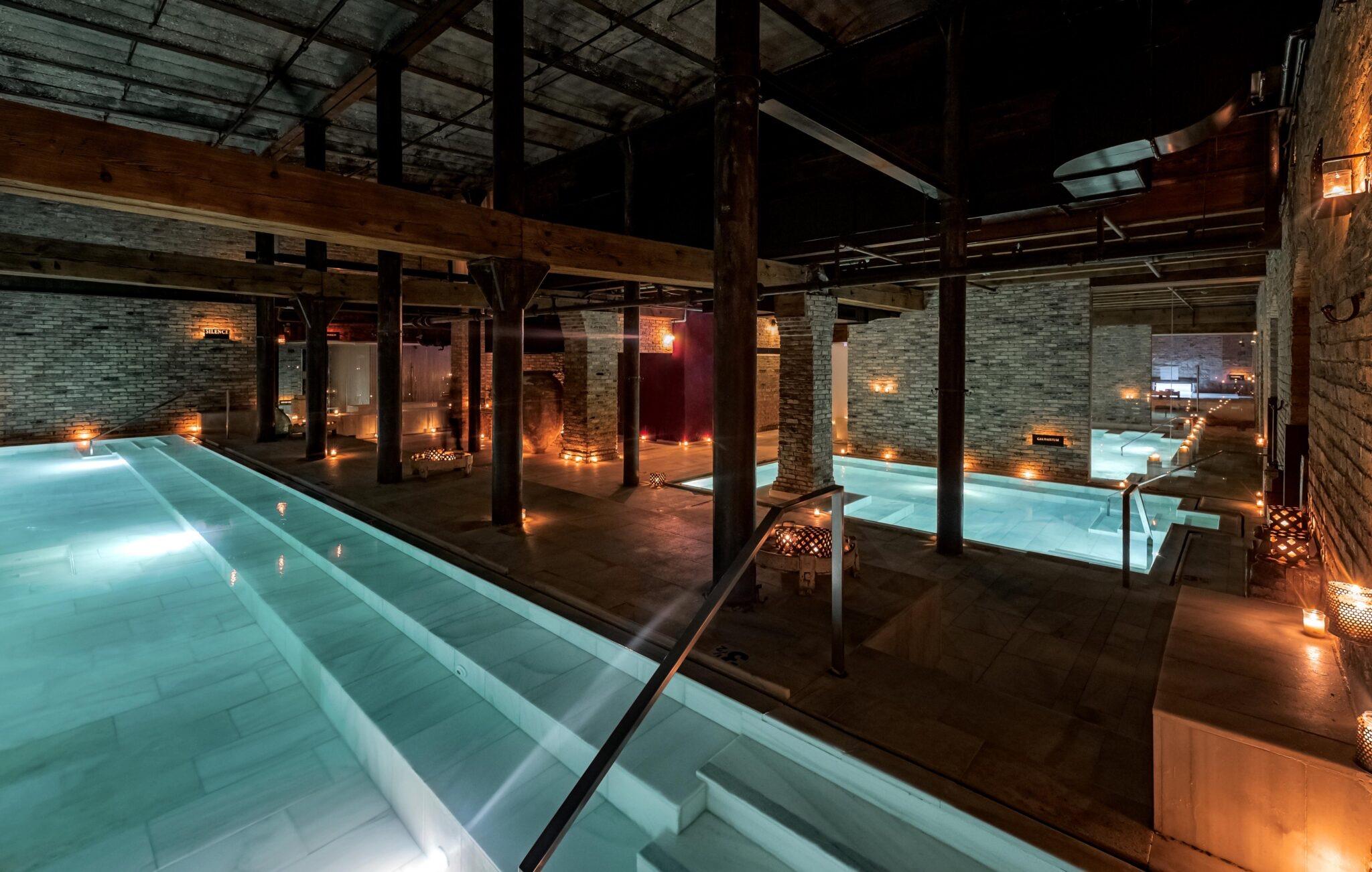 An empty indoor pool under exposed brick arches and timber beams, lit by candles along the water's edge at night.