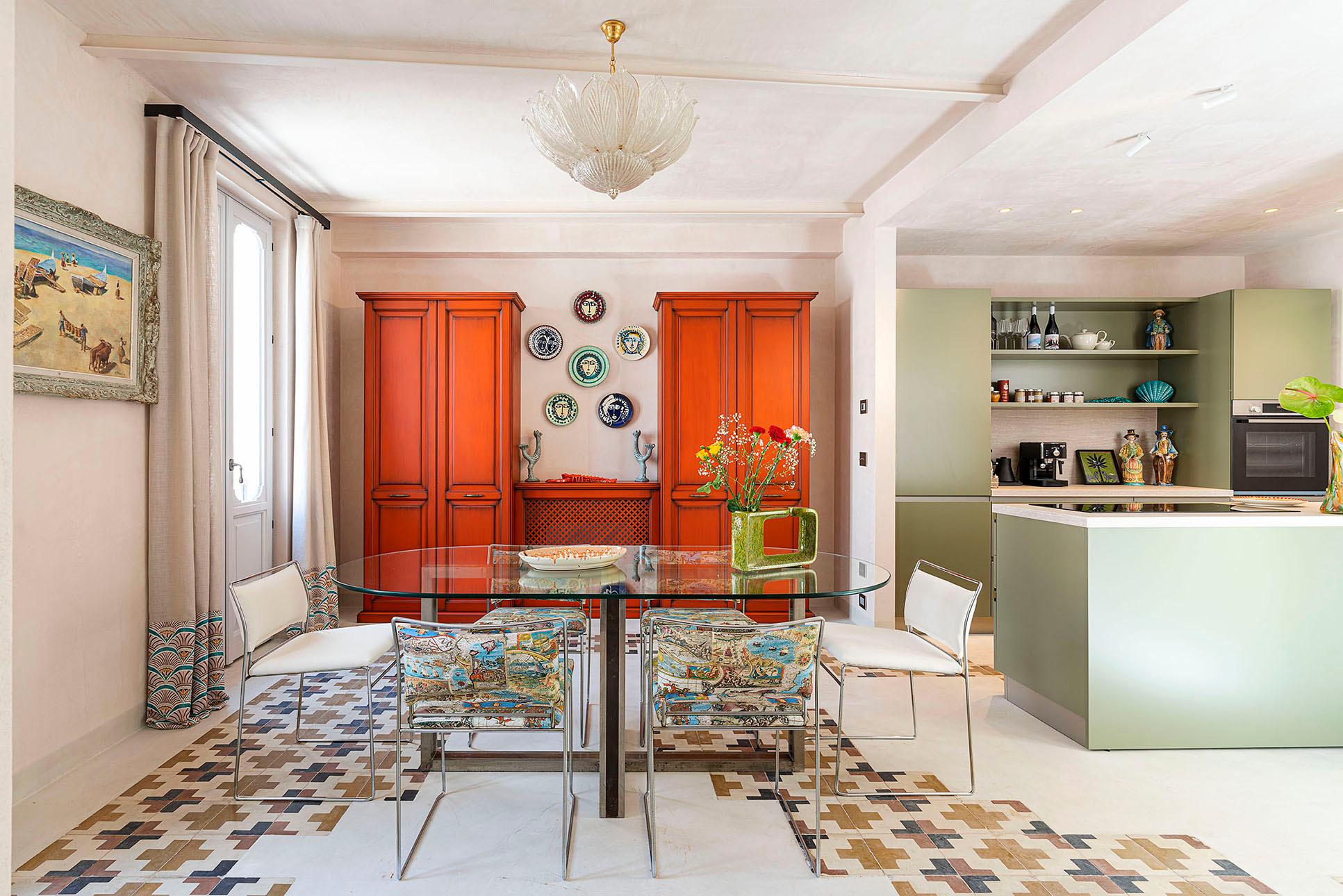 An open plan dining and kitchen area with a glass table and patterned chairs, orange cabinets hung with ceramic plates, and sage green kitchen units beyond.