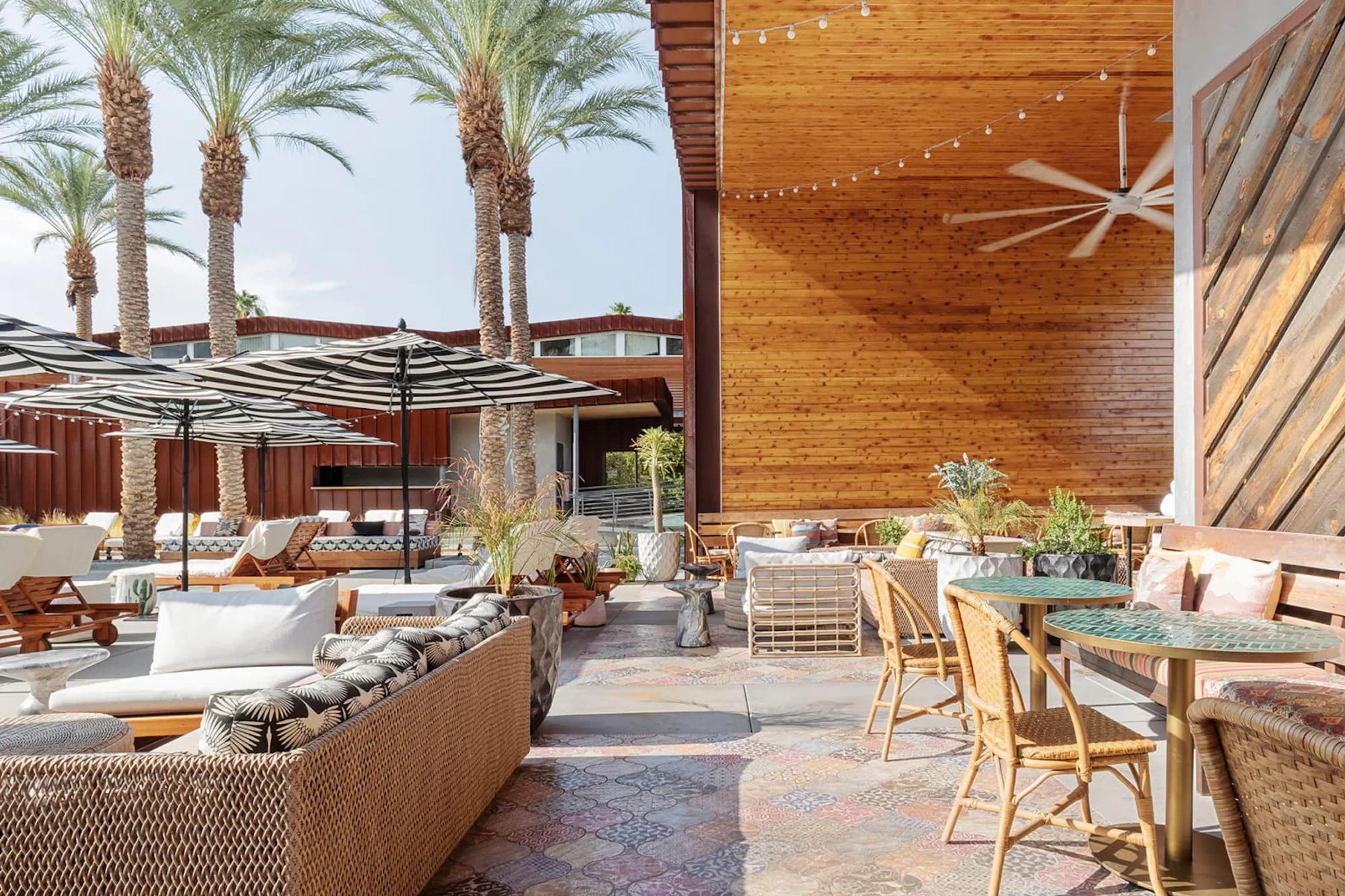 A covered terrace with a timber-clad wall, wicker sofas and cane chairs at round tables with green tops, a ceiling fan and strings of lights overhead, and a planted urn by the seating.