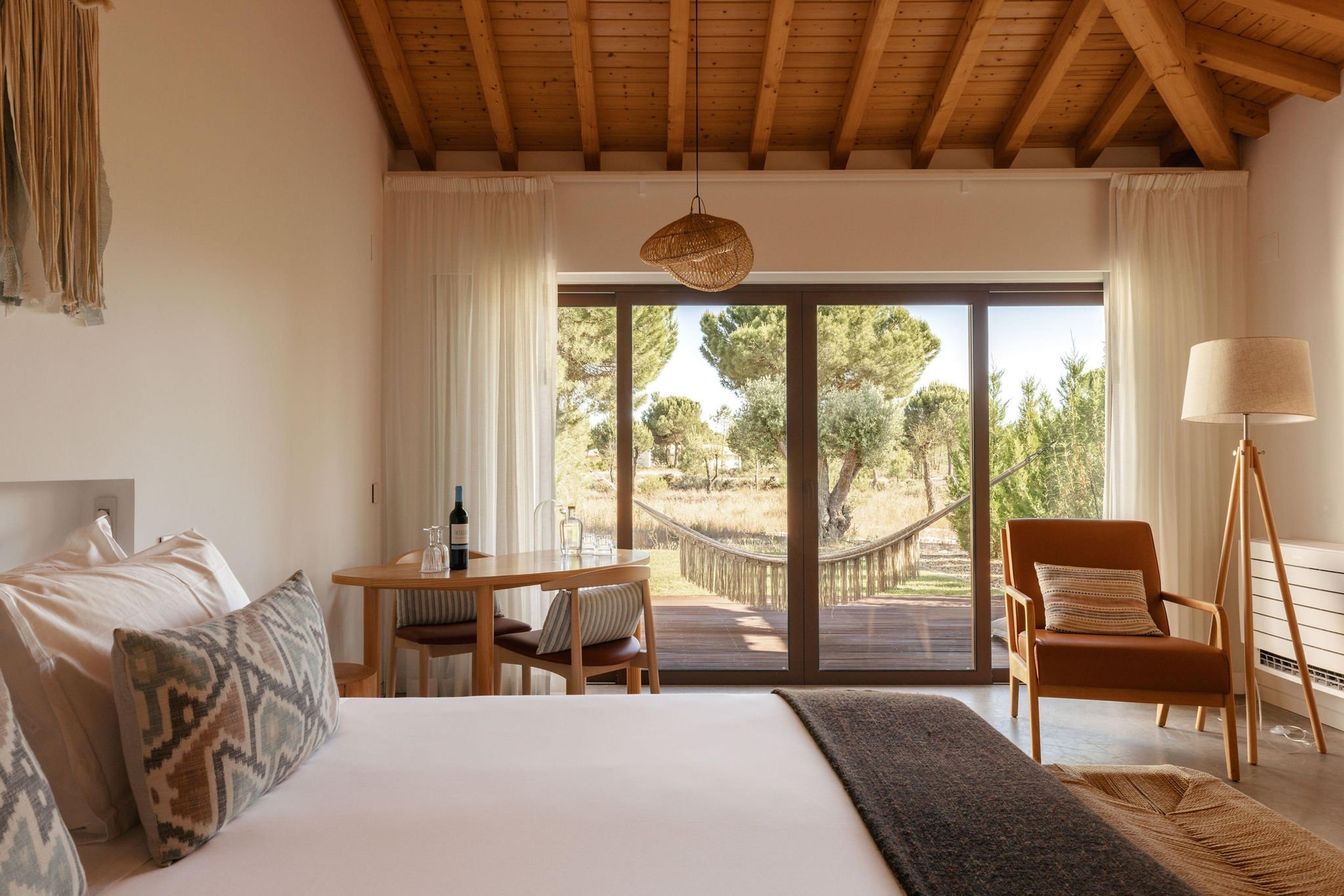 A bedroom with a dining table and wood-beamed ceiling, sliding doors open to a hammock strung between trees outside.