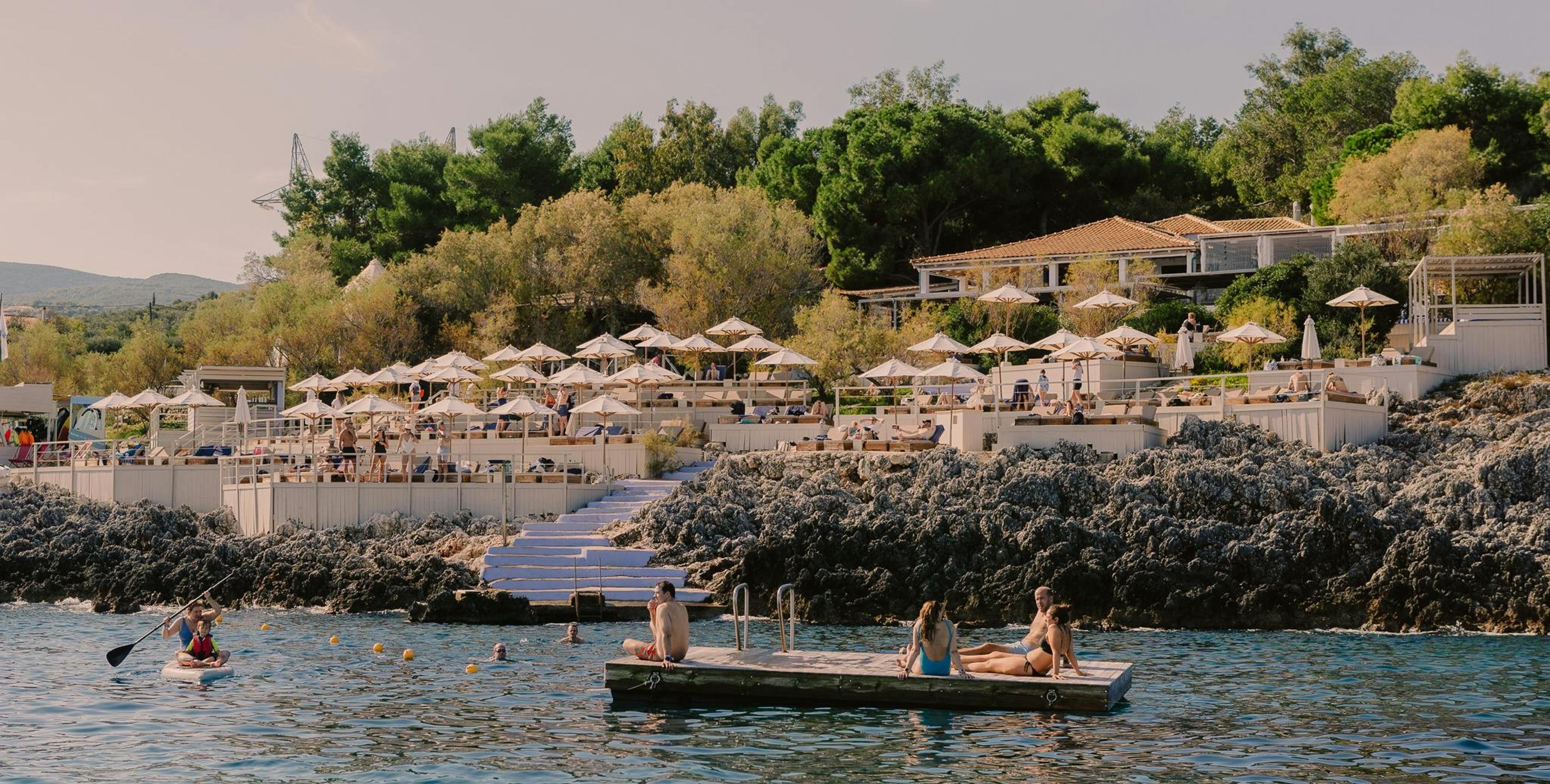 A hillside beach club in evening light, with umbrella seating on a terrace above a rocky shoreline, and people resting on a floating platform and a paddleboard in the sea below.