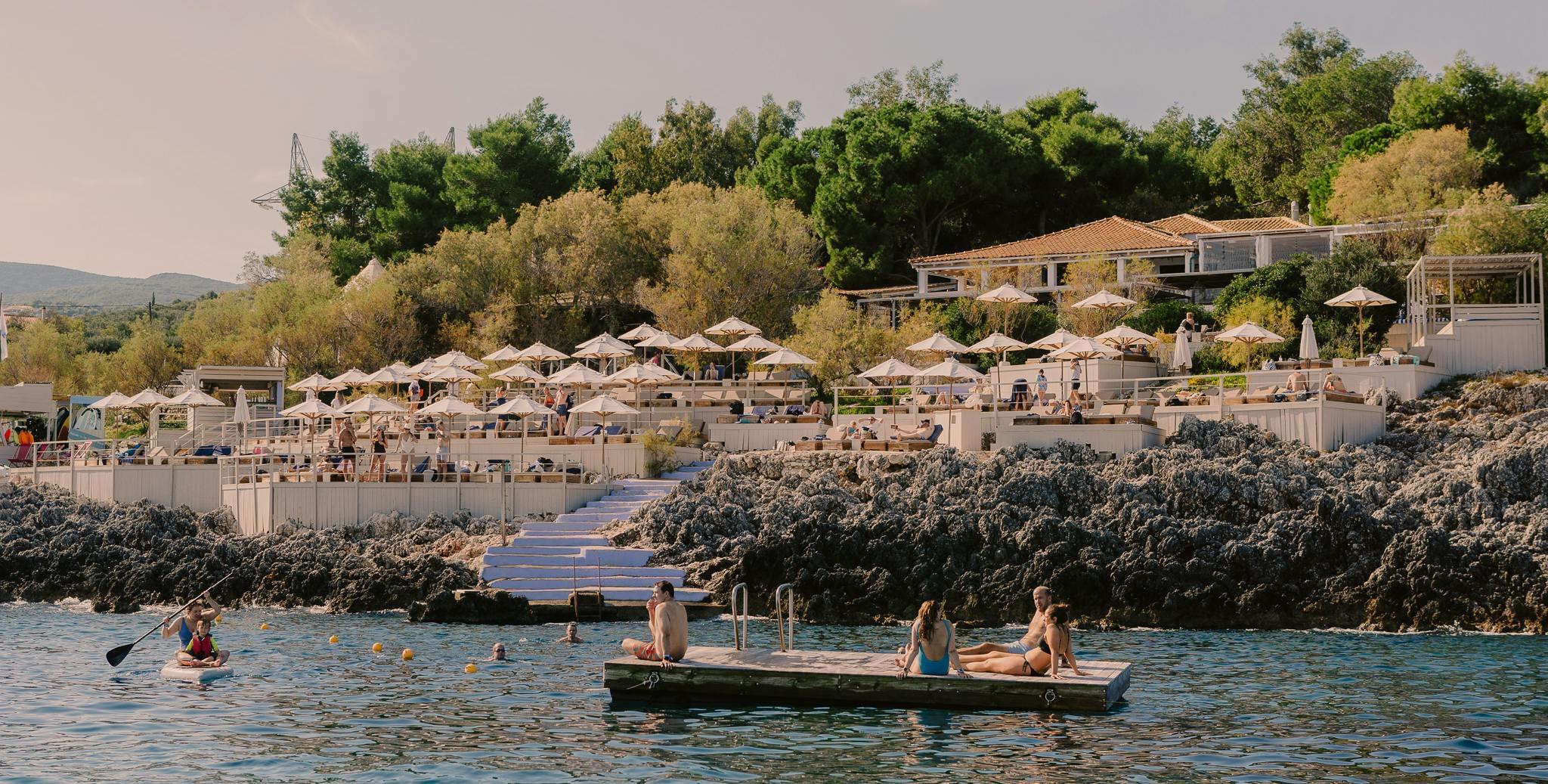 A hillside beach club in evening light, with umbrella seating on a terrace above a rocky shoreline, and people resting on a floating platform and a paddleboard in the sea below.