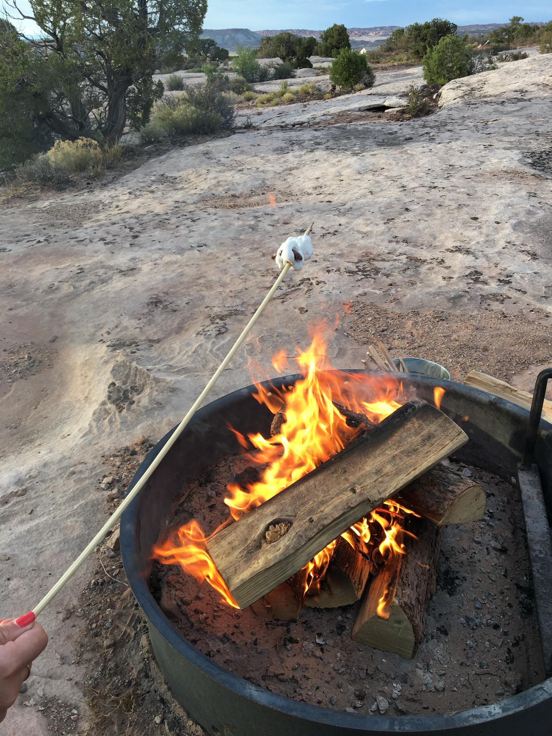 A marshmallow on a stick held over a log fire burning in a metal fire pit on red rock