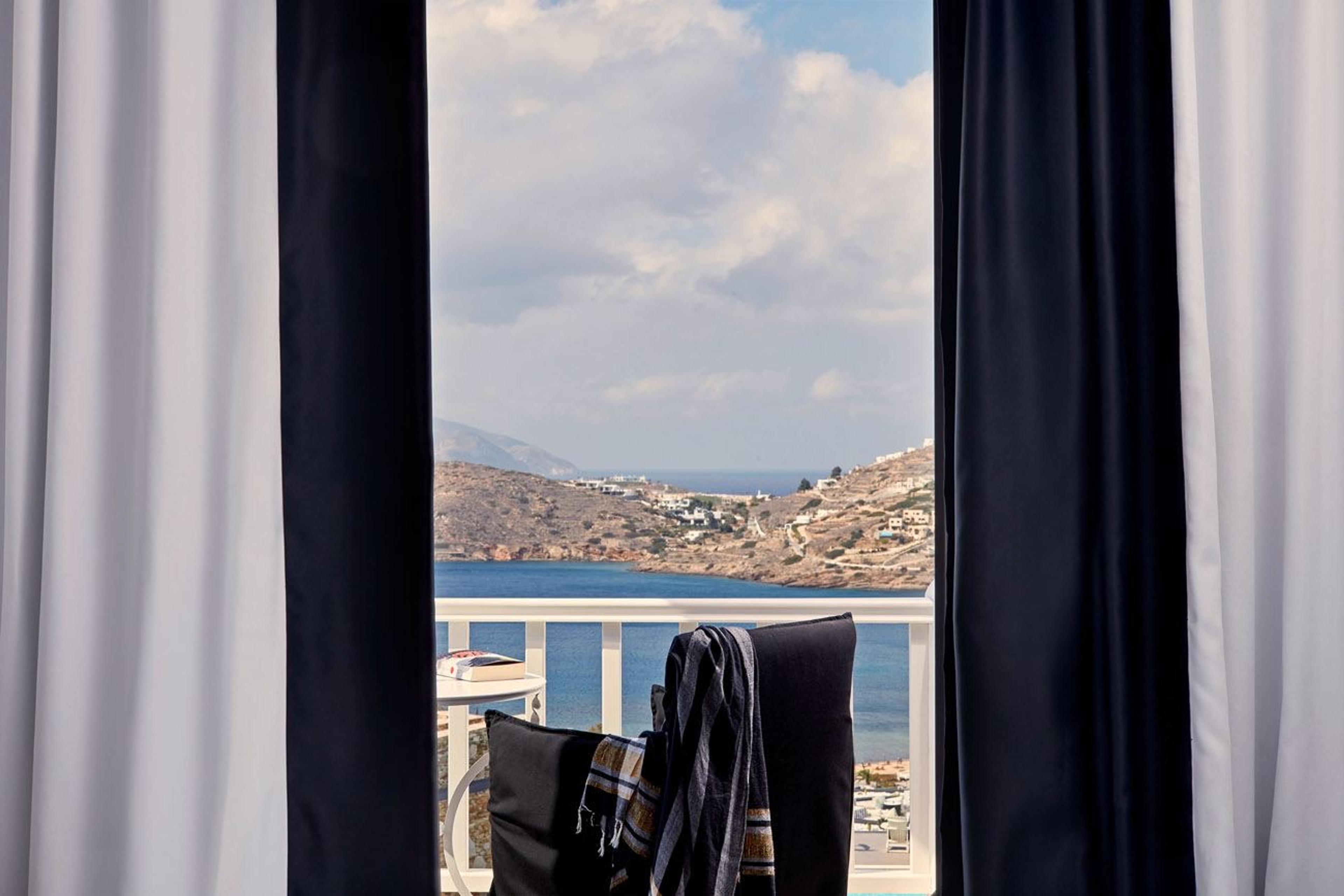 Through open dark and white curtains, a balcony with a folded striped towel, a small table and a book looks over a bay with a white village on the far slope.