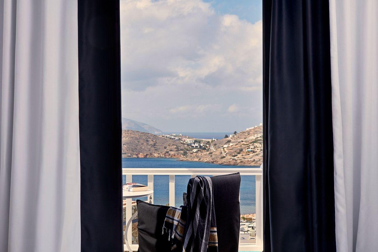 Through open dark and white curtains, a balcony with a folded striped towel, a small table and a book looks over a bay with a white village on the far slope.