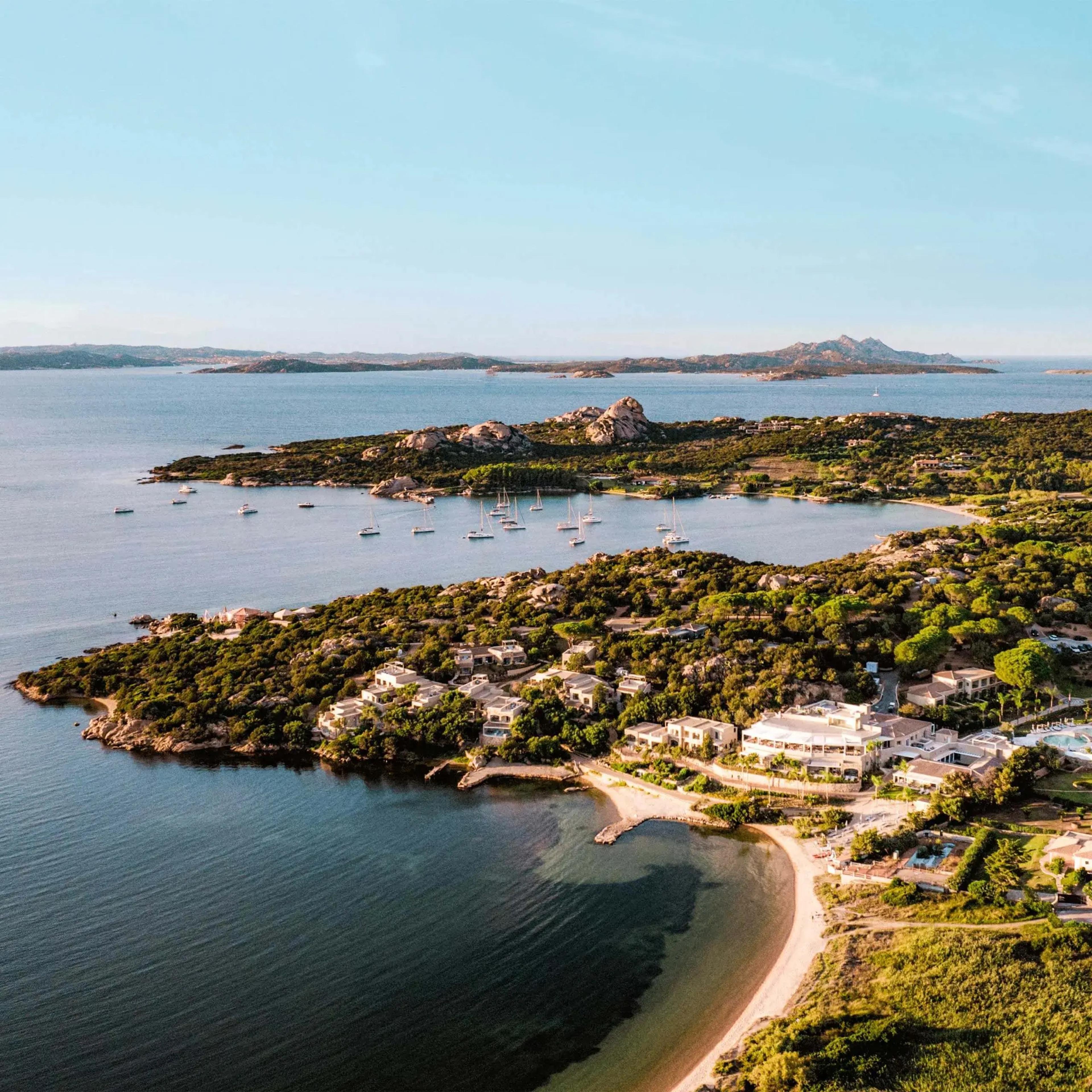 An aerial view of a sheltered bay with anchored sailboats, a rocky pine covered headland, resort buildings along the shore, and distant islands.