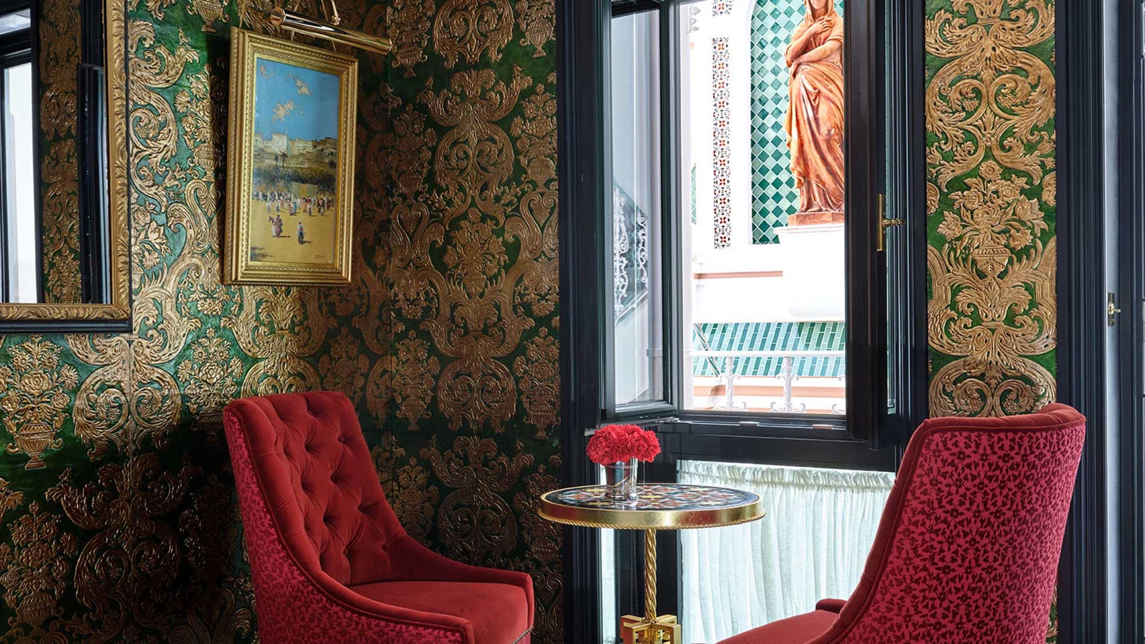 Two red armchairs by an open window with a round table, looking out onto a tiled balcony and a terracotta statue.