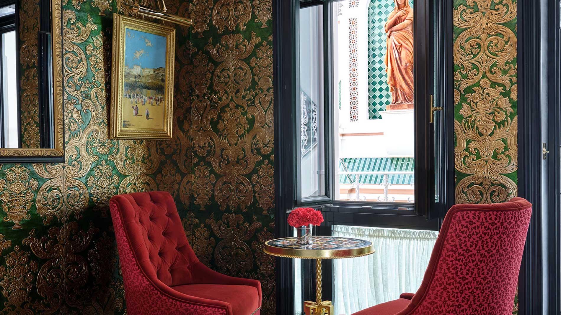 Two red armchairs by an open window with a round table, looking out onto a tiled balcony and a terracotta statue.
