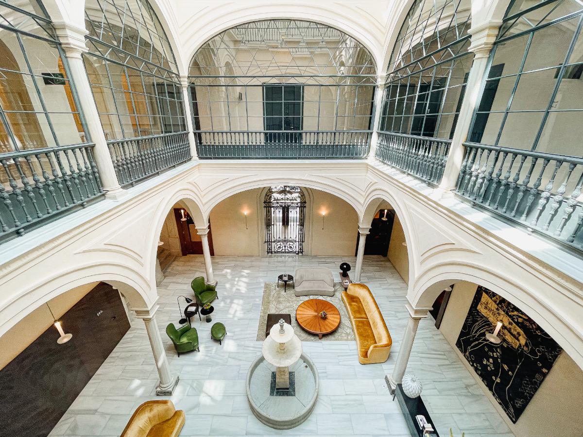 Seen from an upper gallery, a glass-roofed courtyard holds a marble fountain, gold velvet sofas, and green armchairs, ringed by arched walkways on two floors.