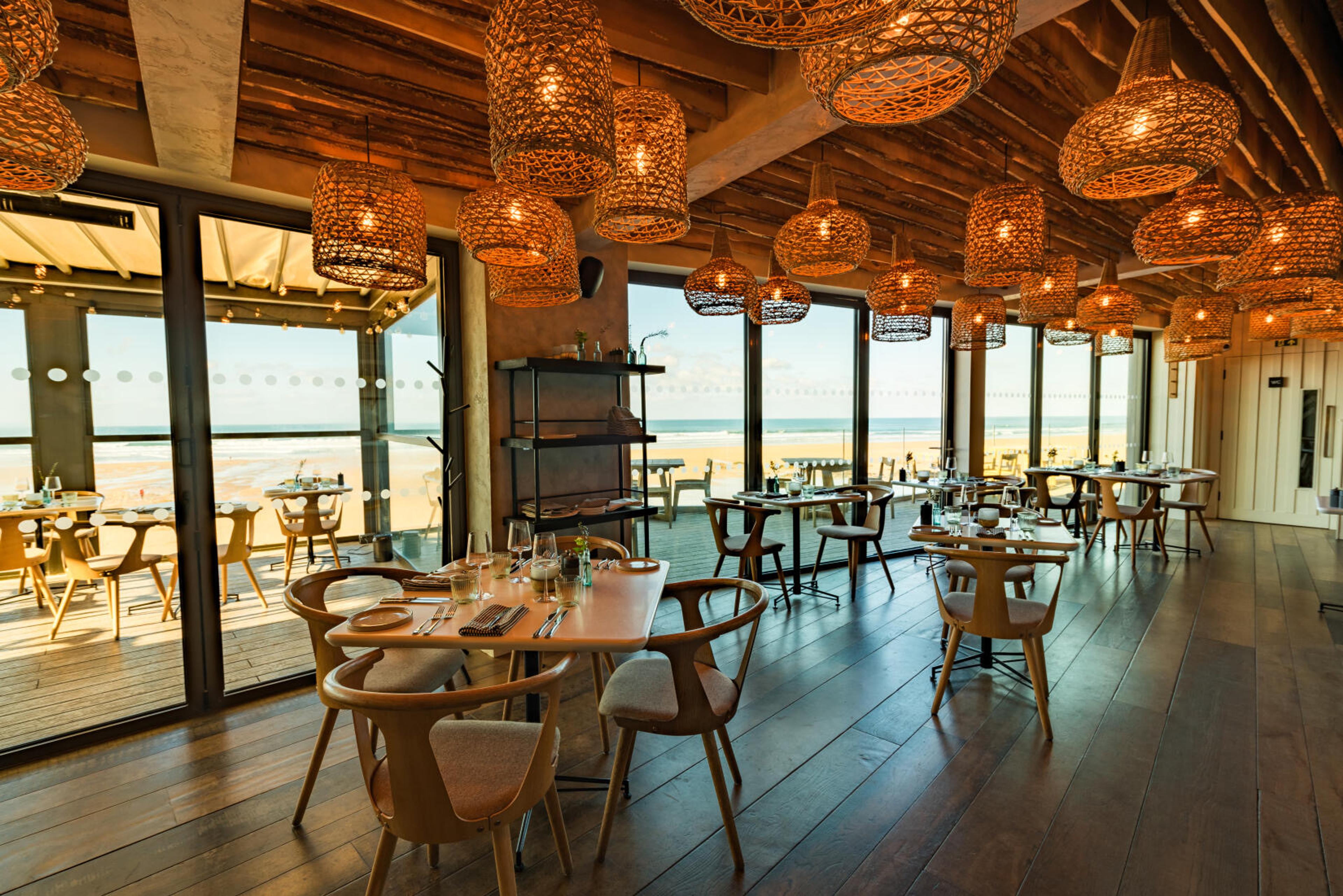 A restaurant dining room with woven pendant lights over set tables and glass doors open to the beach.