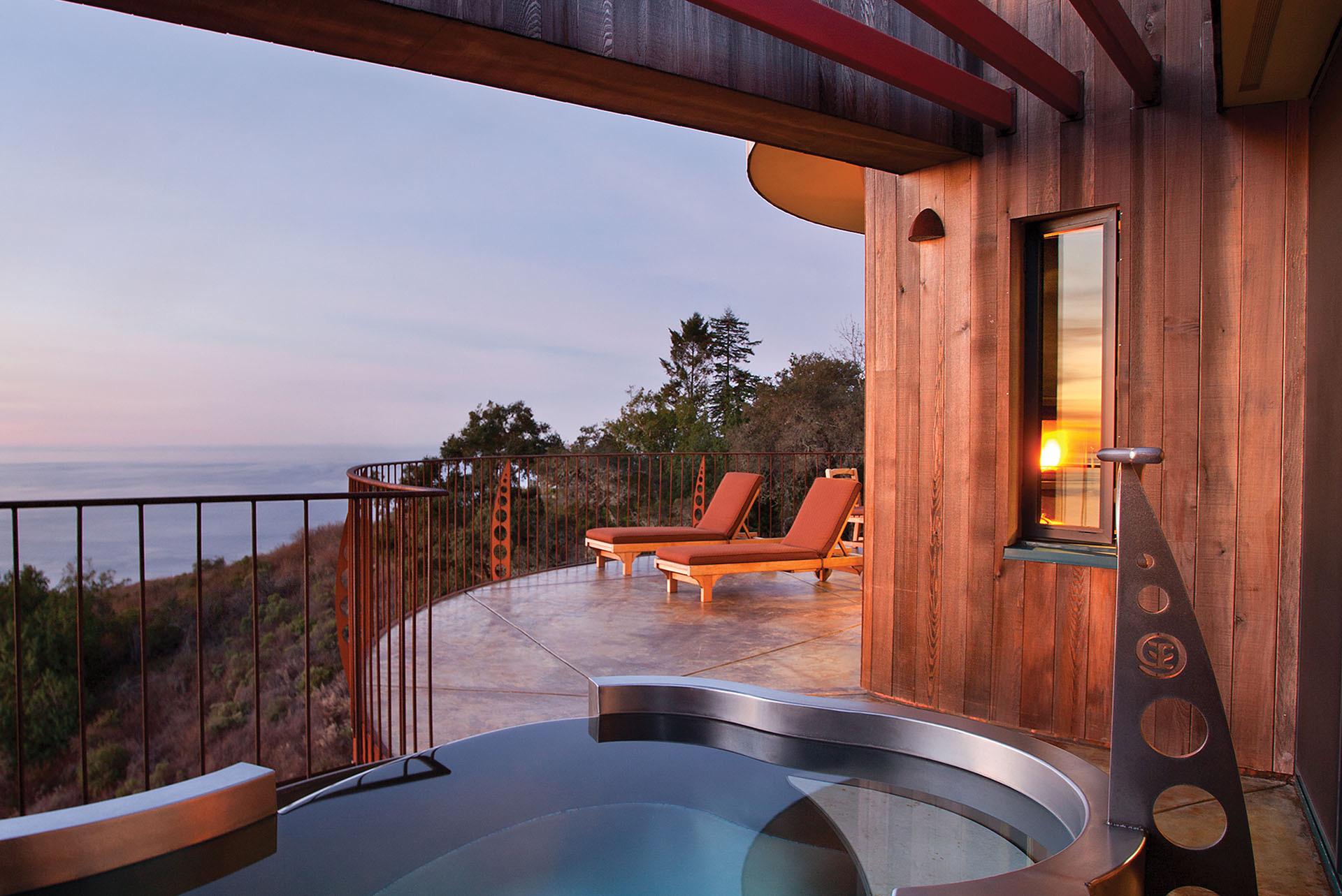 A round stainless hot tub on a wooden deck beside two patio loungers, with a wood-paneled guest house and the ocean beyond at dusk.