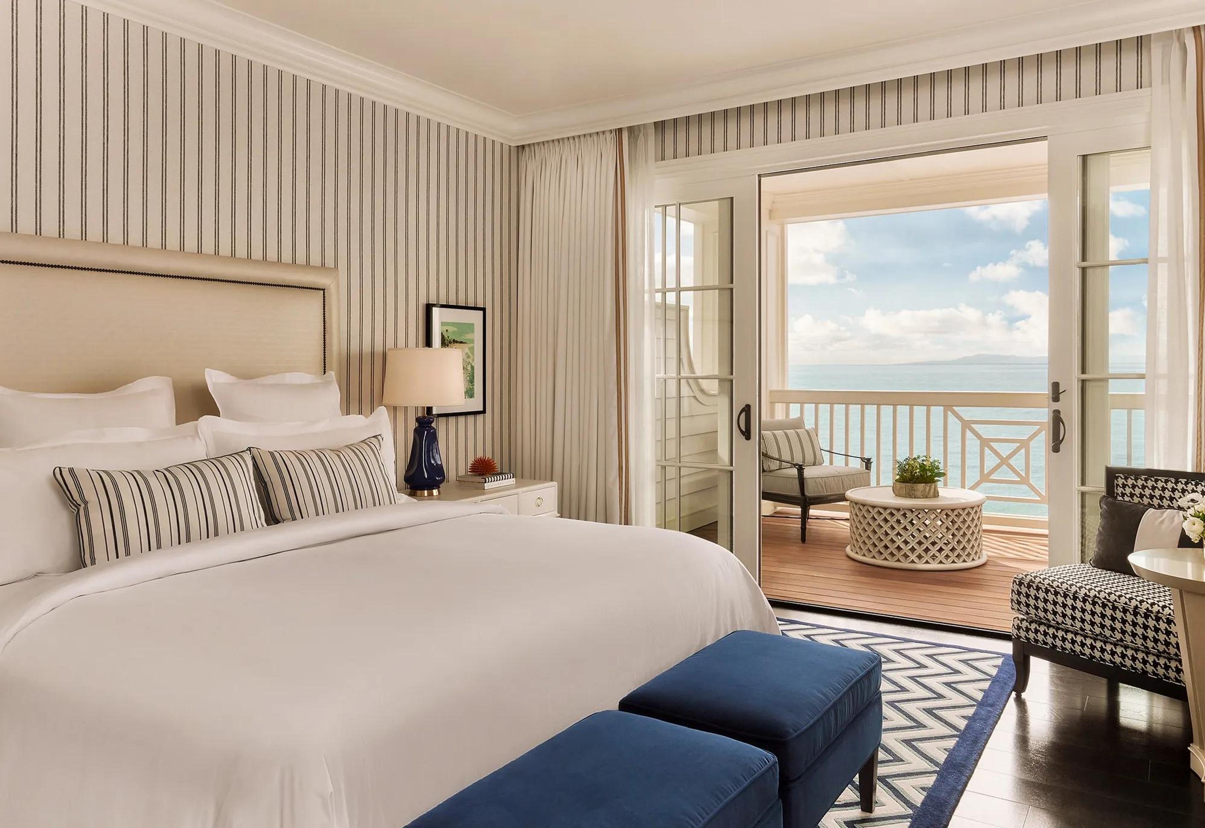 A hotel bedroom with a white upholstered headboard and a blue bench, its balcony doors open to the ocean.