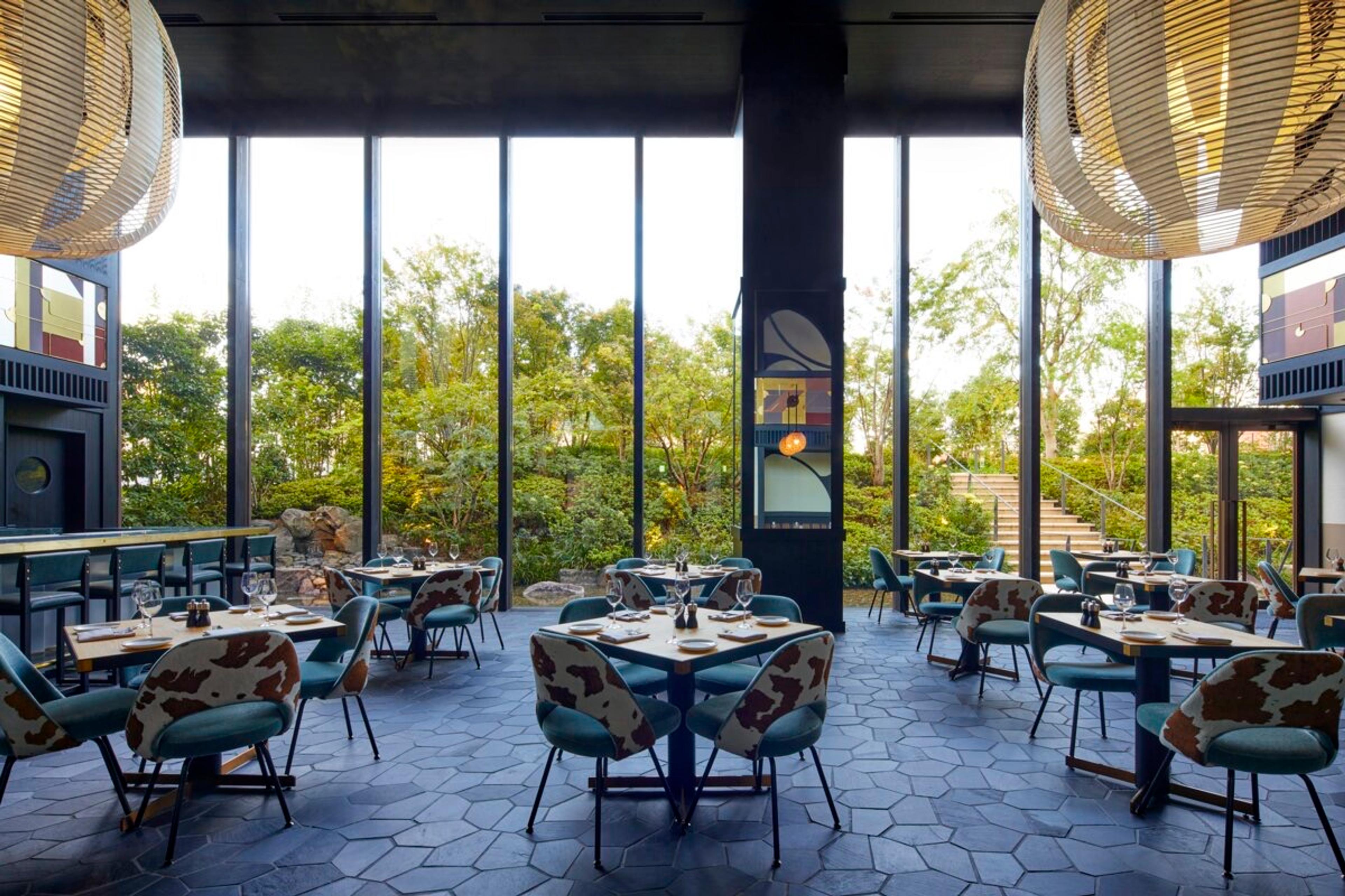 A restaurant dining room with cow print upholstered chairs and floor to ceiling windows looking onto a garden of trees and a staircase.
