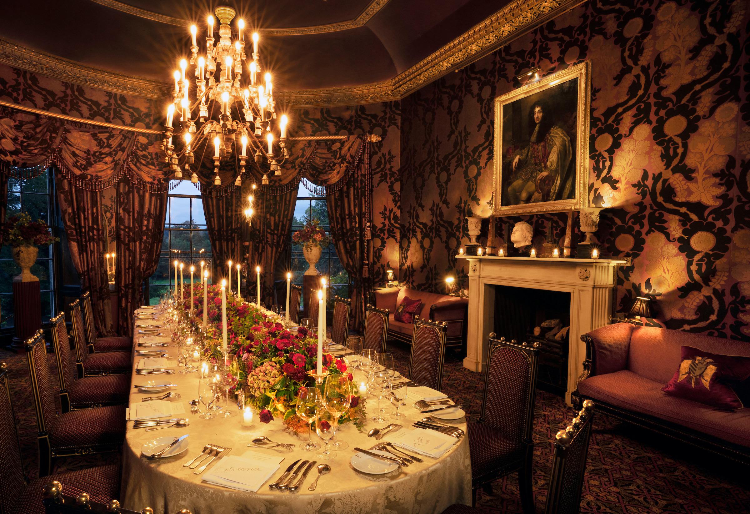 an oval table laid for a formal dinner under a lit chandelier, in a room with patterned walls, a fireplace and a portrait