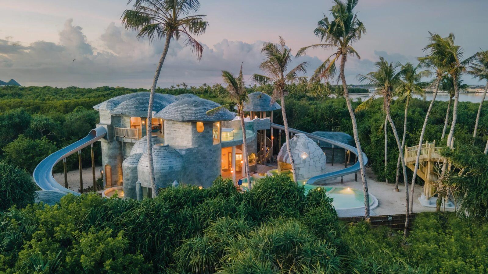 an overwater villa with dome shaped thatched roofs and a blue slide curving into a pool, lit at dusk and surrounded by palm trees
