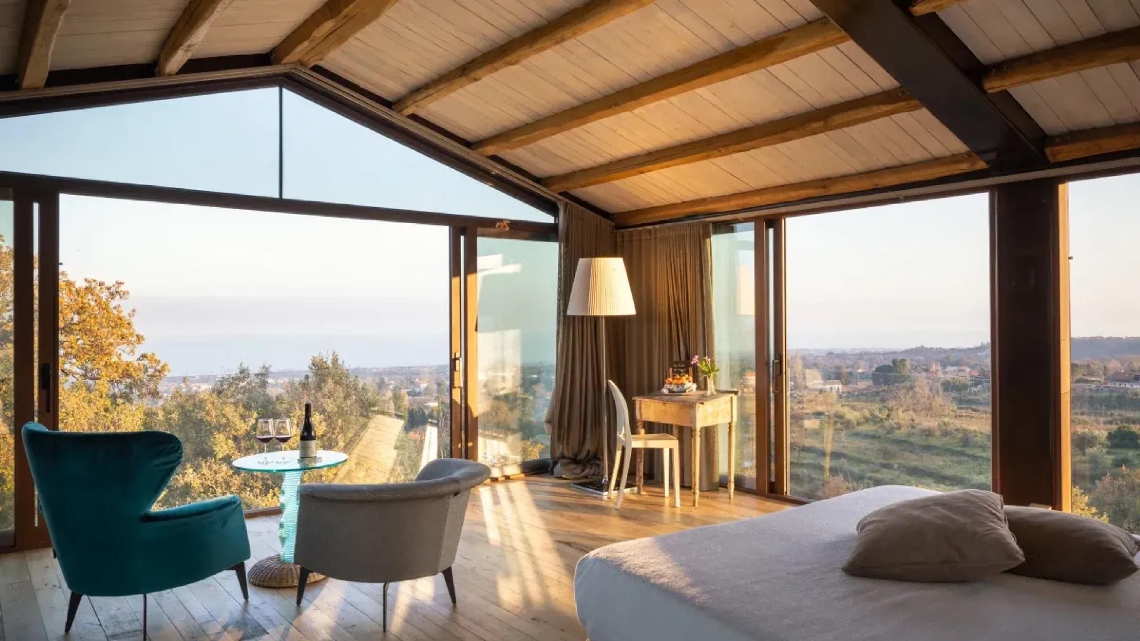 A bedroom opening on two sides to floor to ceiling glass, with two armchairs and a round table set with wine, overlooking hills and the sea.