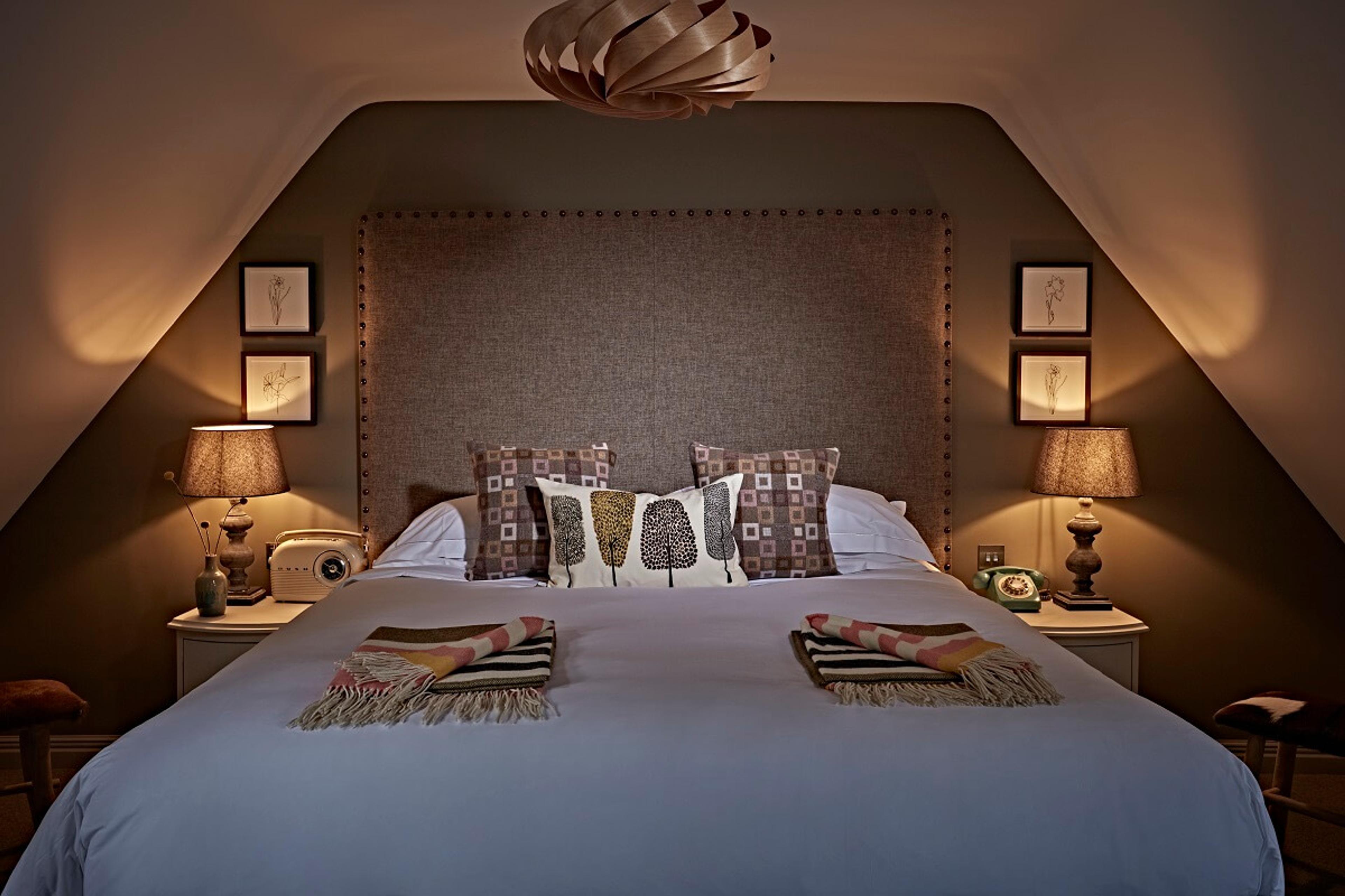 A snug bedroom tucked under eaves, with a buttoned headboard, two amber bedside lamps and folded tartan throws laid across the end of the bed.