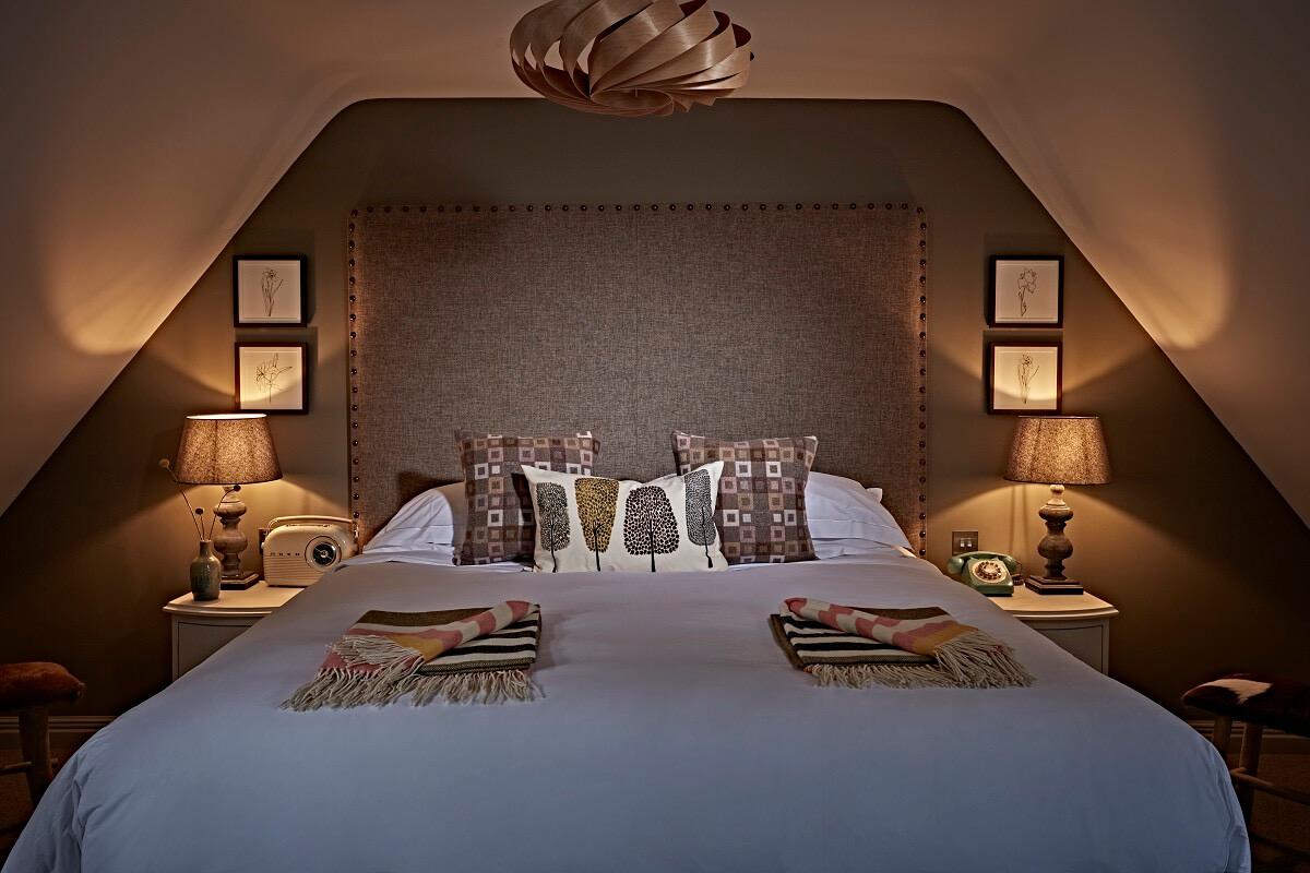 A snug bedroom tucked under eaves, with a buttoned headboard, two amber bedside lamps and folded tartan throws laid across the end of the bed.