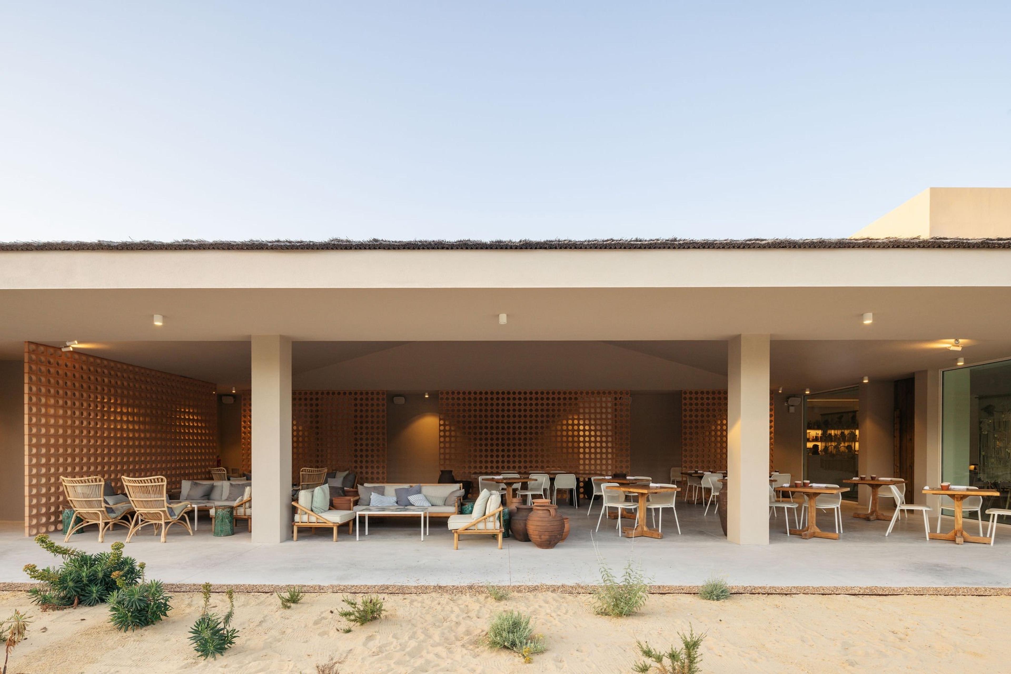 A covered terrace with patterned screen walls, rattan armchairs, a sofa and dining tables set with chairs, large ceramic jars on the floor.