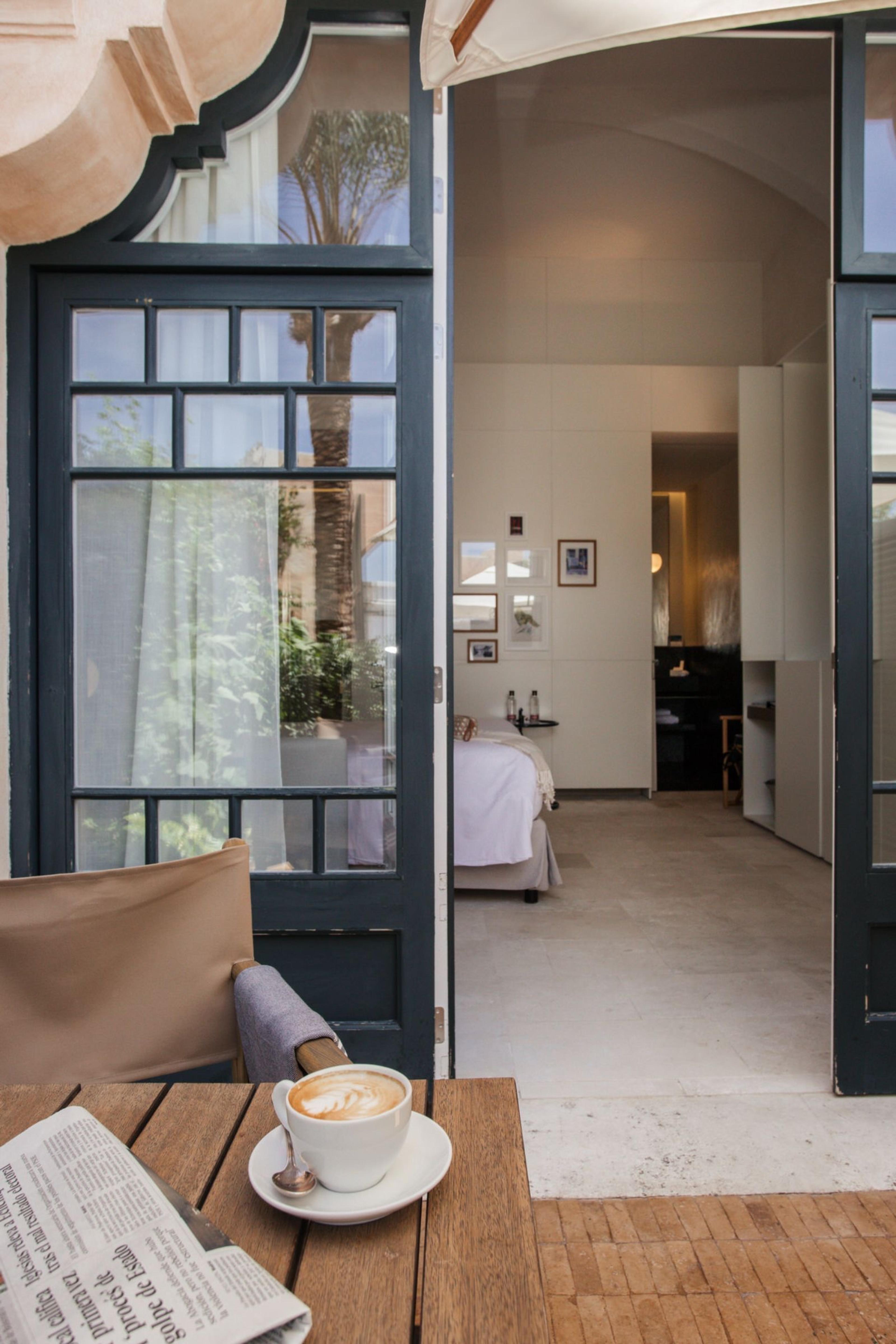 A coffee cup and a folded newspaper on an outdoor table seen through open glazed doors, with a bedroom visible beyond.