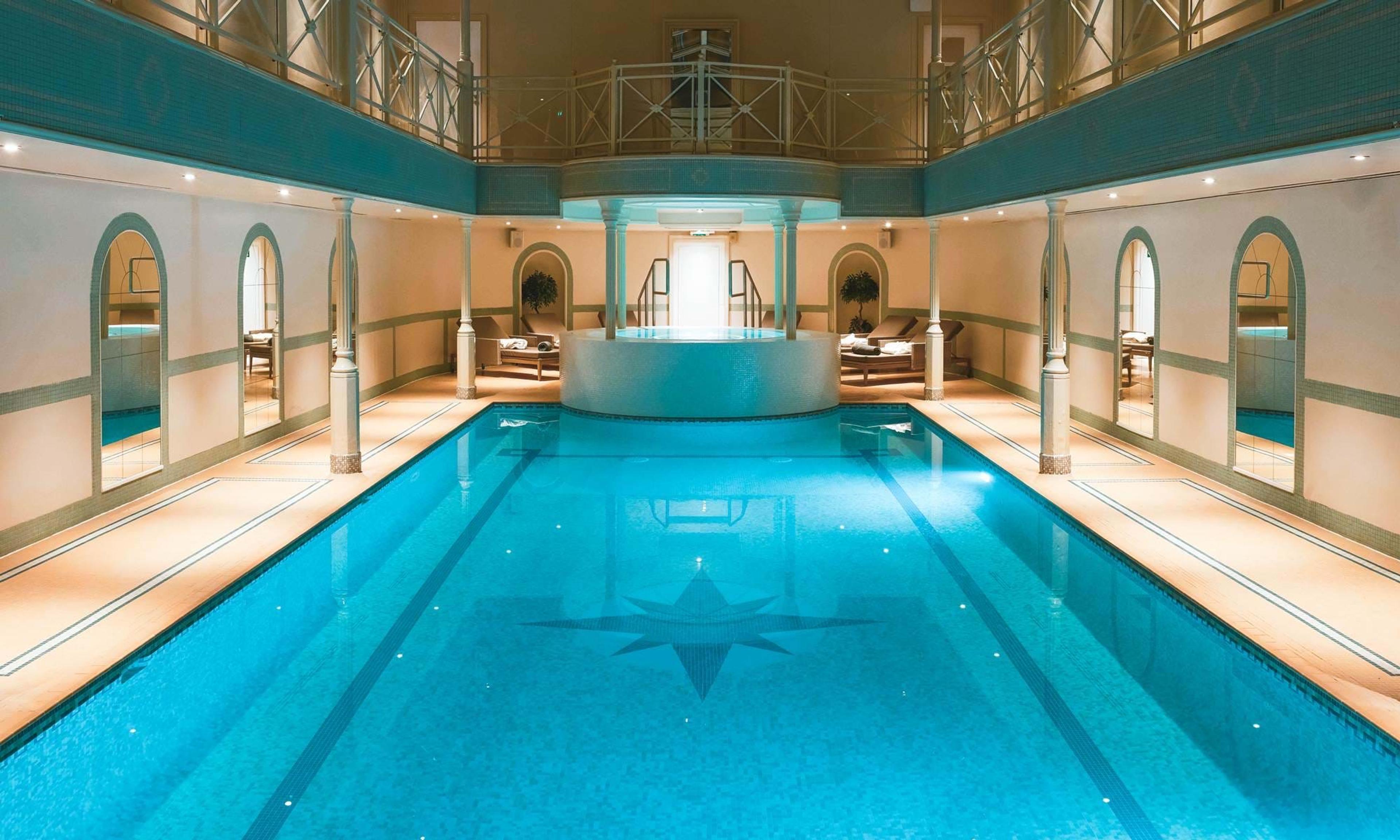 An empty indoor swimming pool lined with arched alcoves and loungers, under a two-storey balustraded gallery.