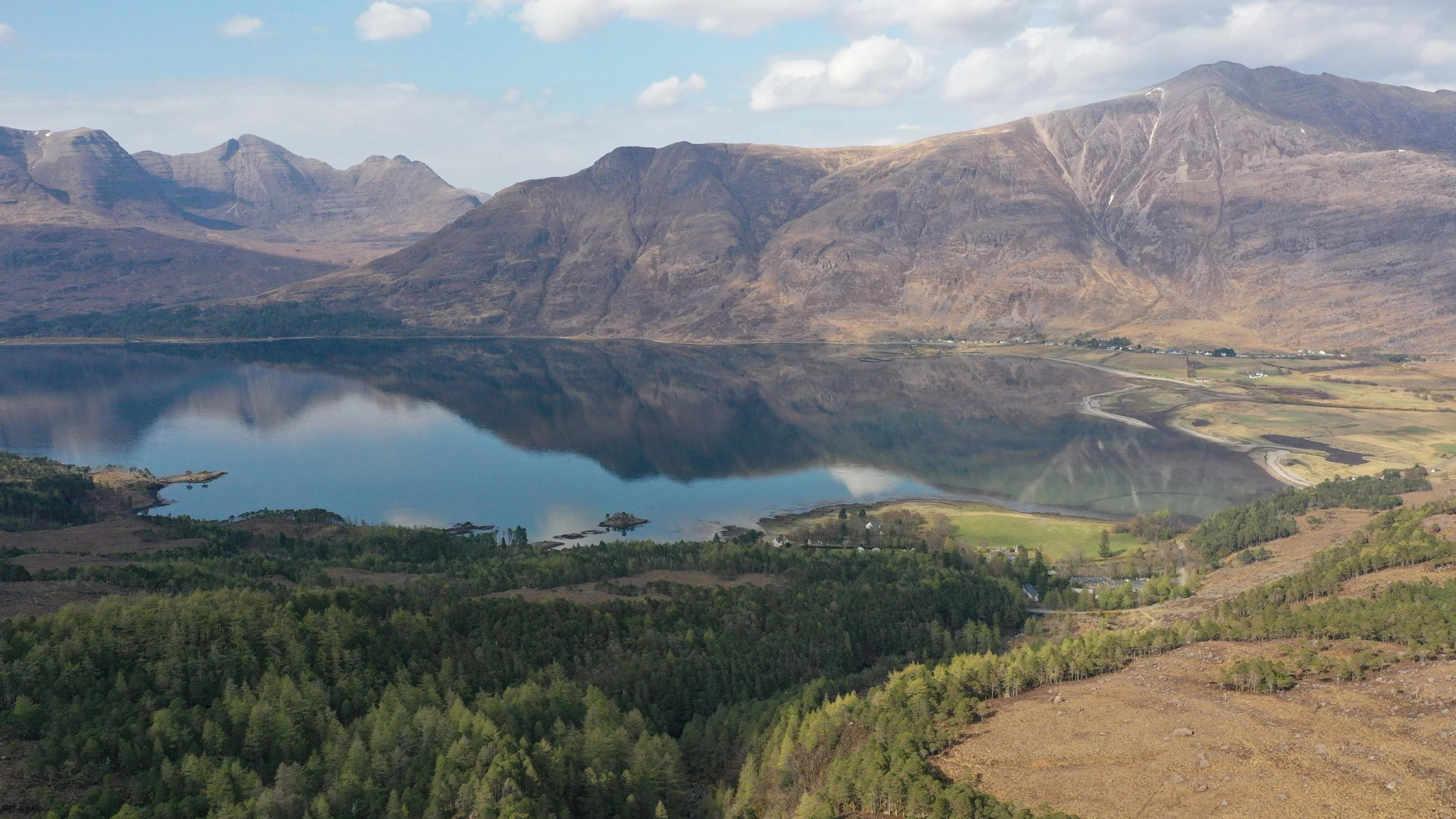 An aerial view of a loch bordered by forest and bare mountains, the still water reflecting the peaks around it.