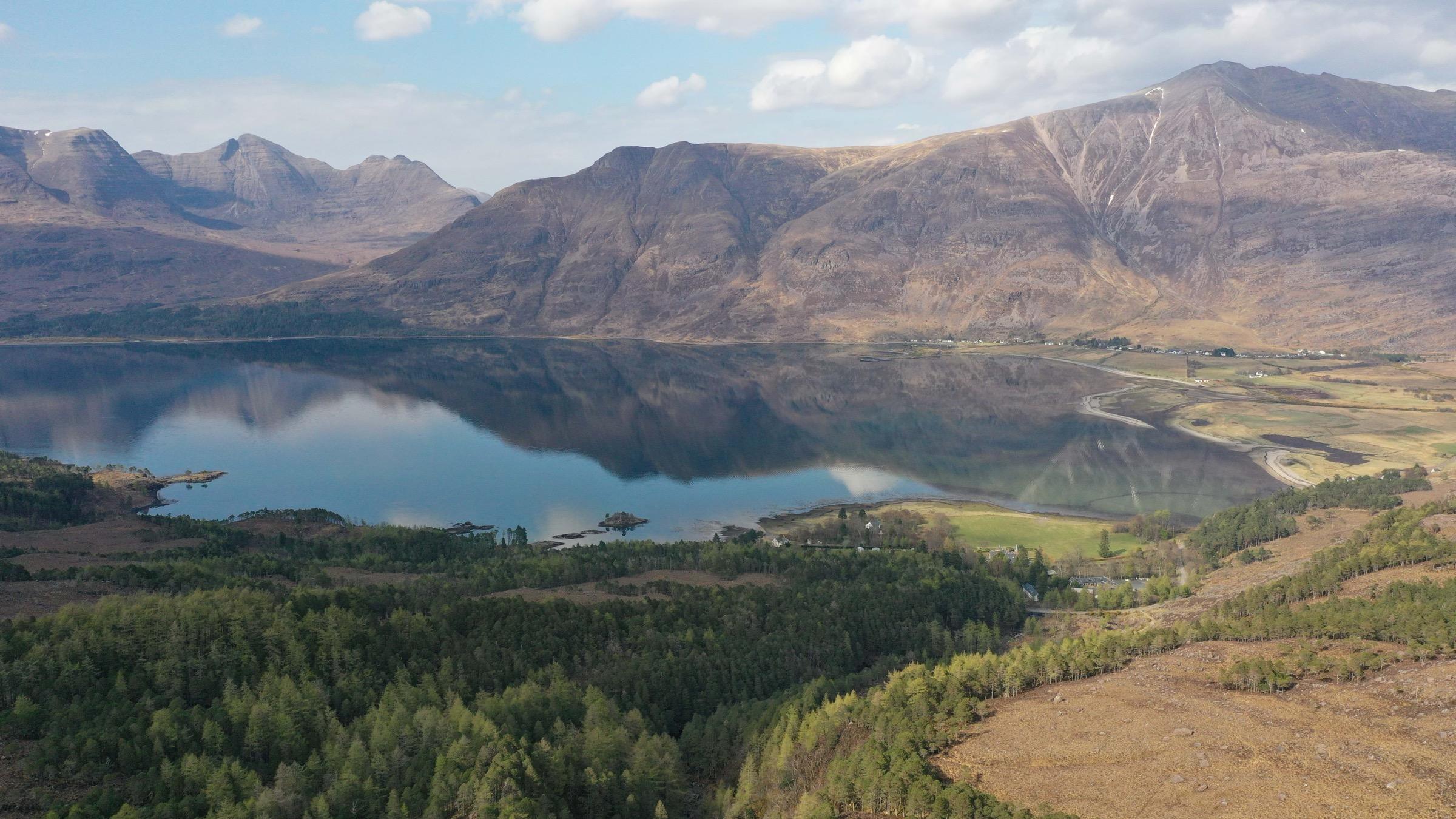 An aerial view of a loch bordered by forest and bare mountains, the still water reflecting the peaks around it.