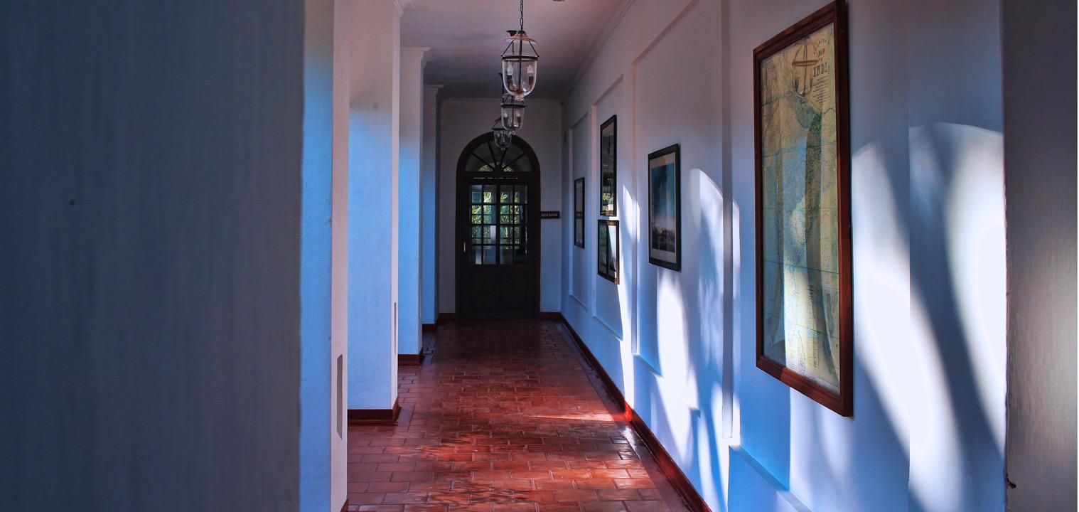 A quiet hallway with framed pictures on one wall, a red tiled floor, and an arched wooden door at the far end lit by hanging lanterns.
