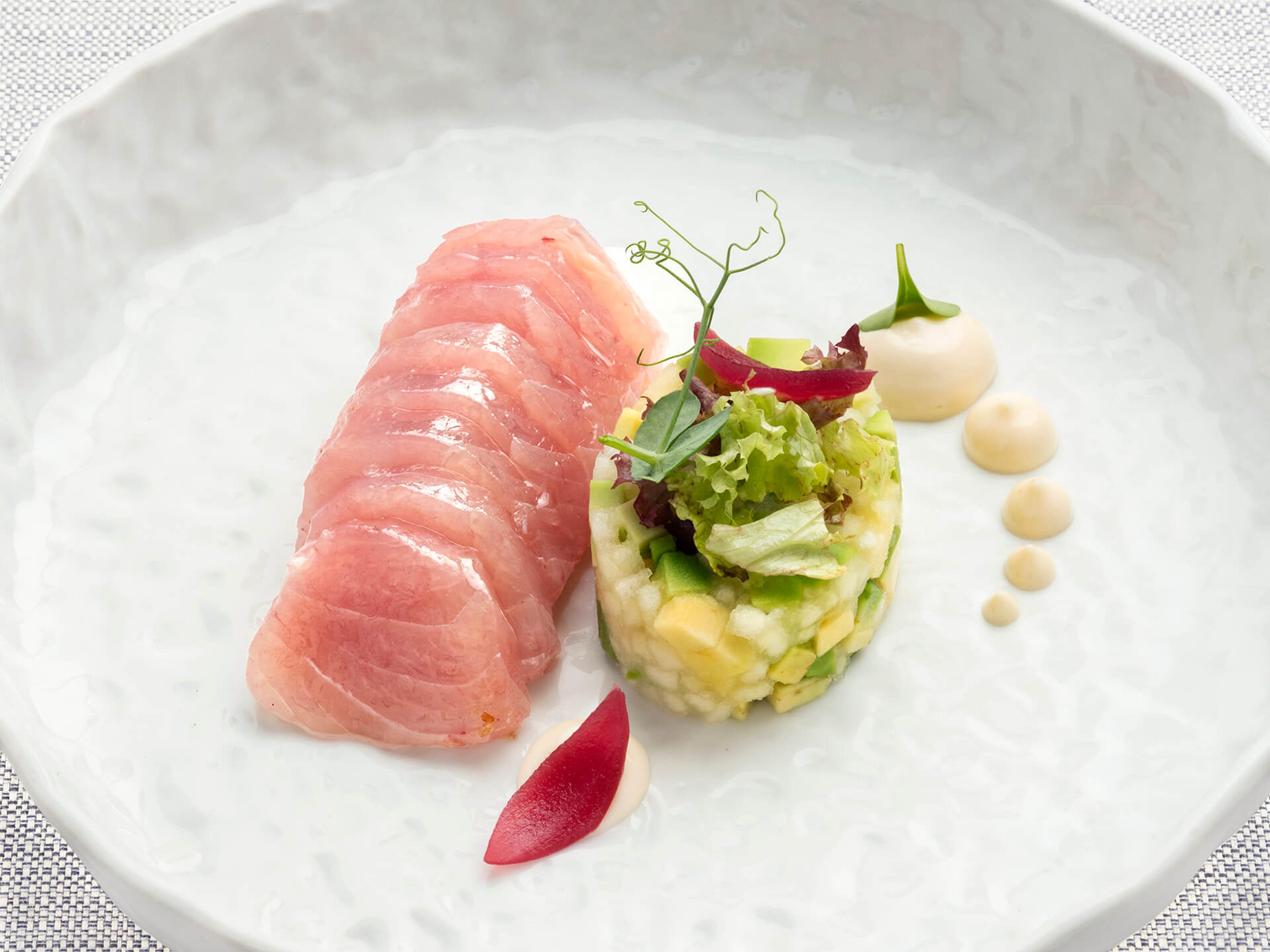 Sliced tuna fanned on a white plate beside a round mound of rice salad with avocado and yellow fruit, with a dot of sauce and a pink onion petal.
