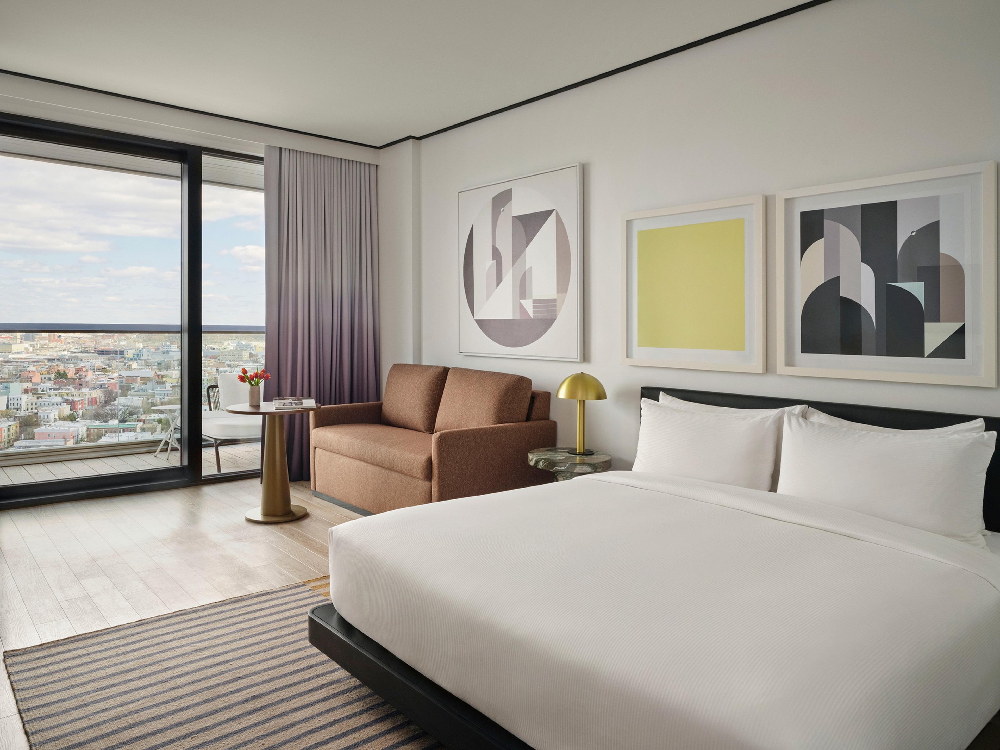A hotel bedroom with a bed, a brown sofa, three abstract artworks, and a floor-to-ceiling window showing the city skyline.