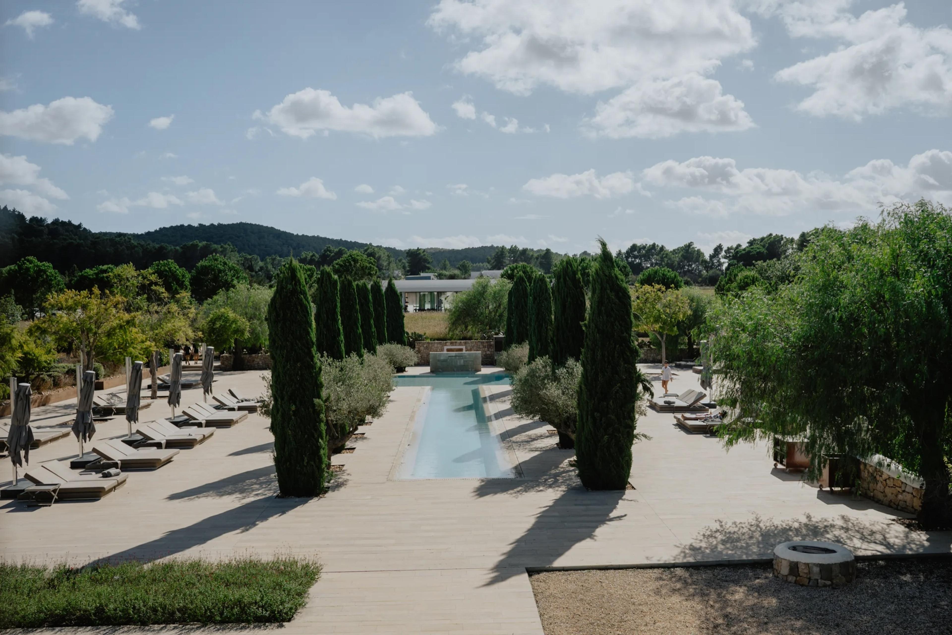 A raised view over a swimming pool lined with cypress trees, sun loungers, and a low building beyond, under a partly cloudy sky.