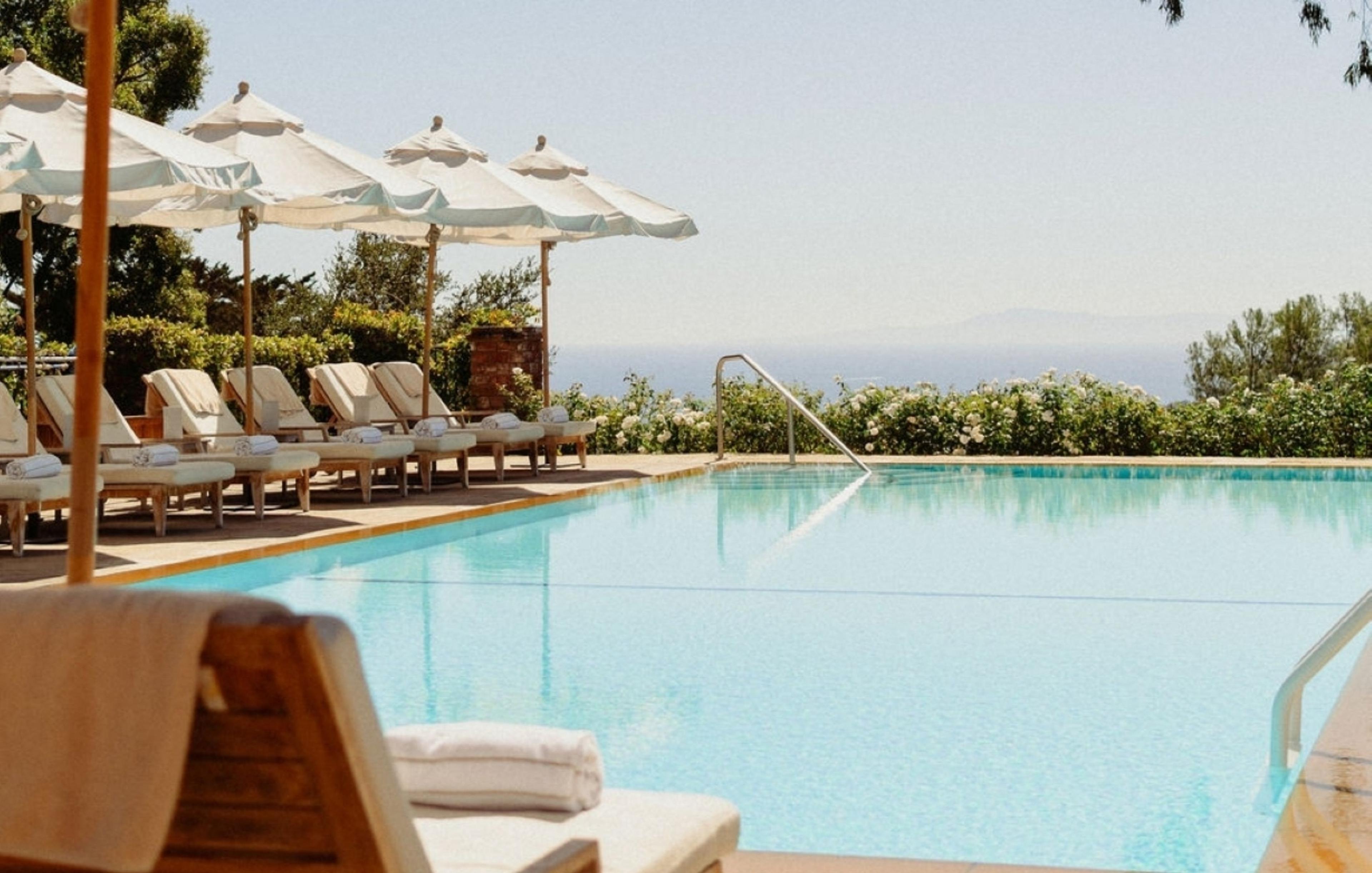 A row of white loungers and umbrellas beside a rectangular pool, with flowering hedges and a hazy stretch of ocean beyond.