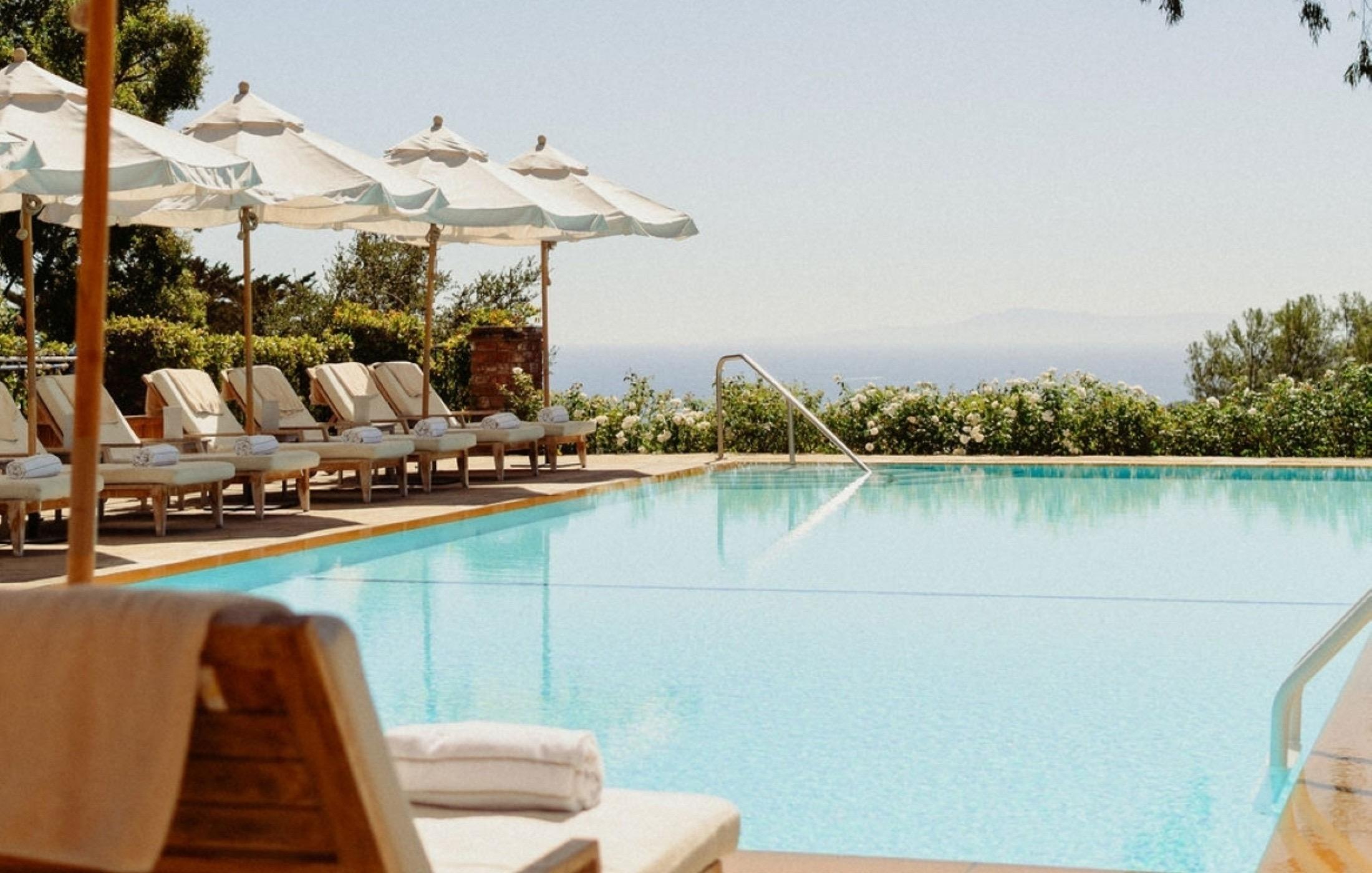 A row of white loungers and umbrellas beside a rectangular pool, with flowering hedges and a hazy stretch of ocean beyond.
