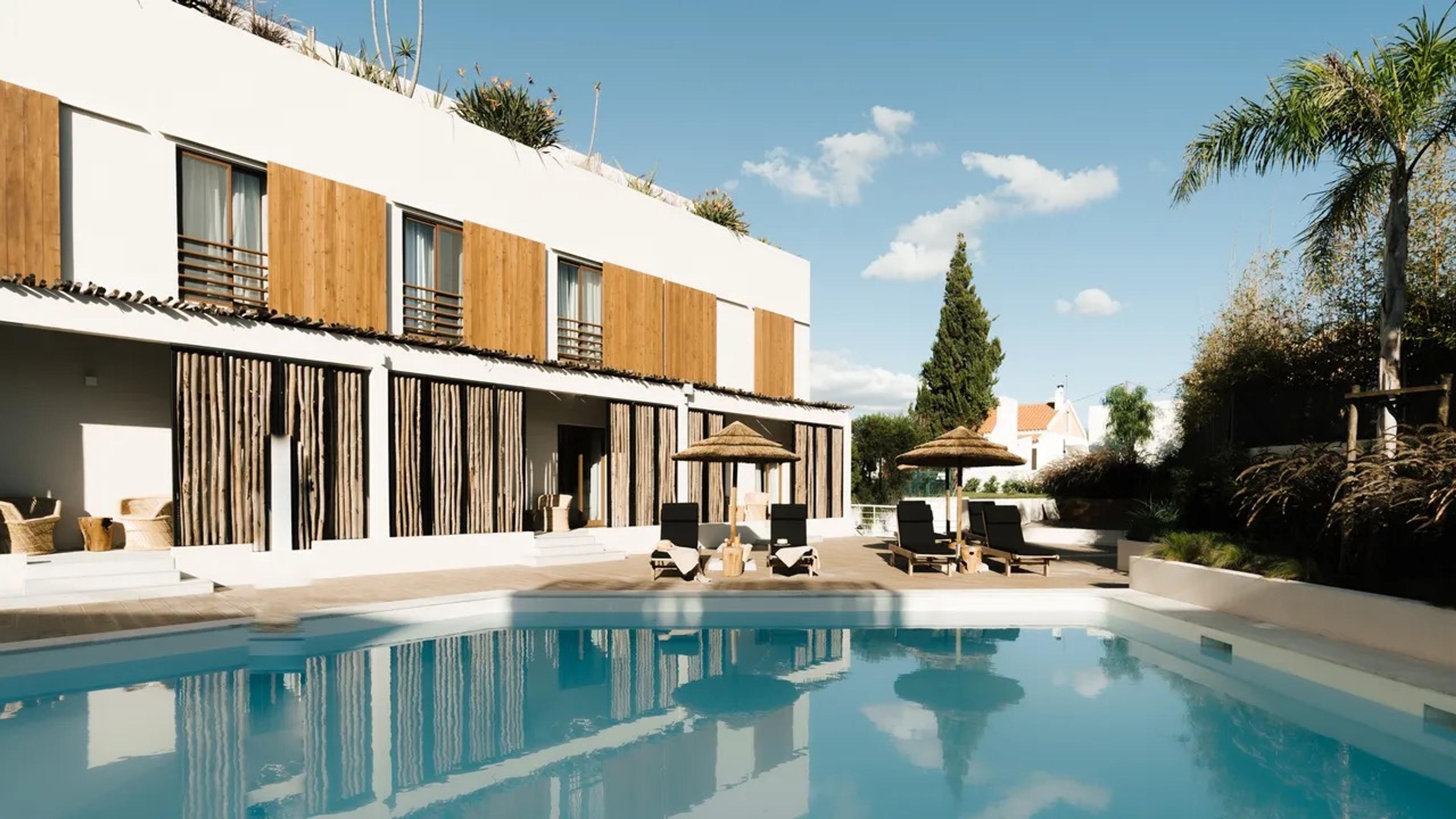 A white building with wood-shuttered windows behind a rectangular pool, with straw parasols and sun loungers on the terrace and palm trees to the side.