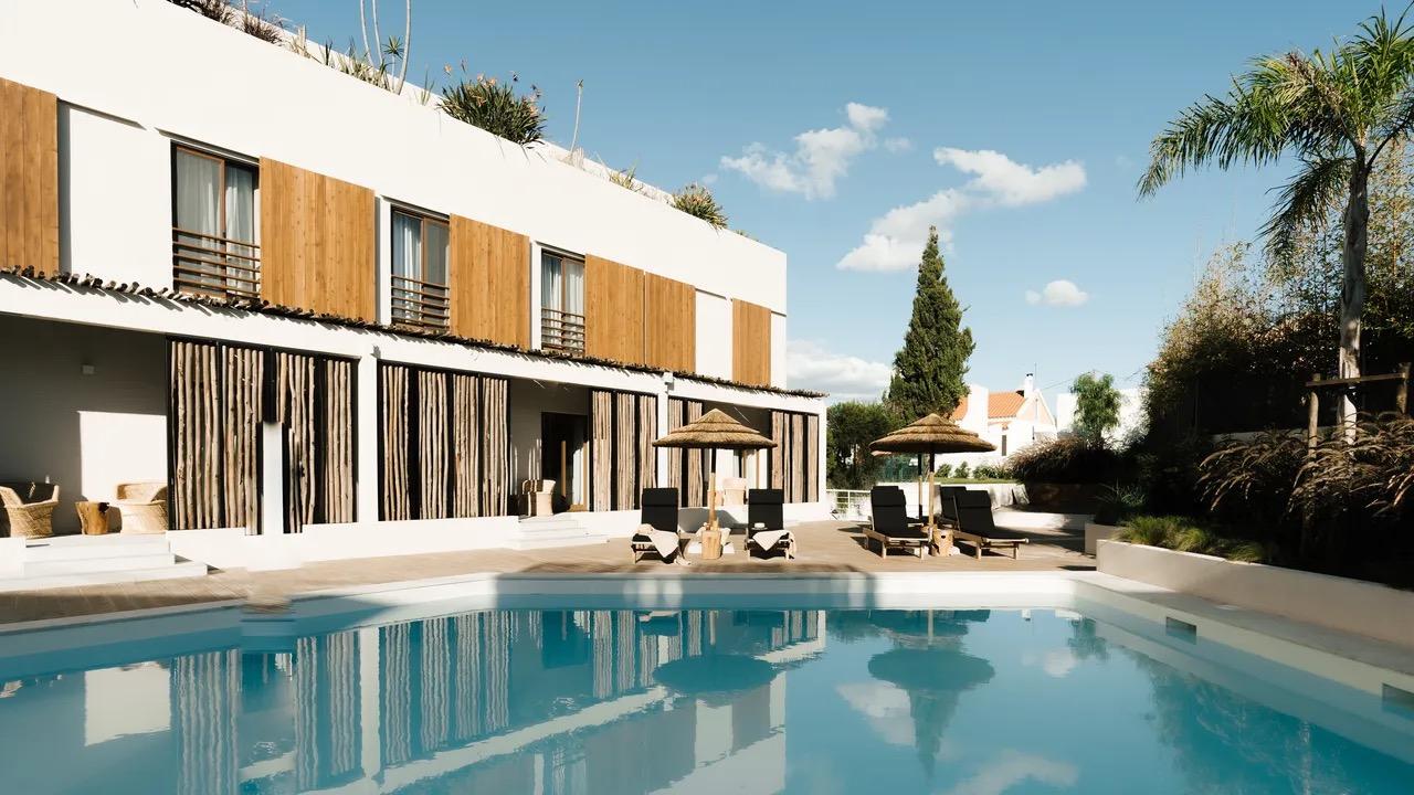 A white building with wood-shuttered windows behind a rectangular pool, with straw parasols and sun loungers on the terrace and palm trees to the side.