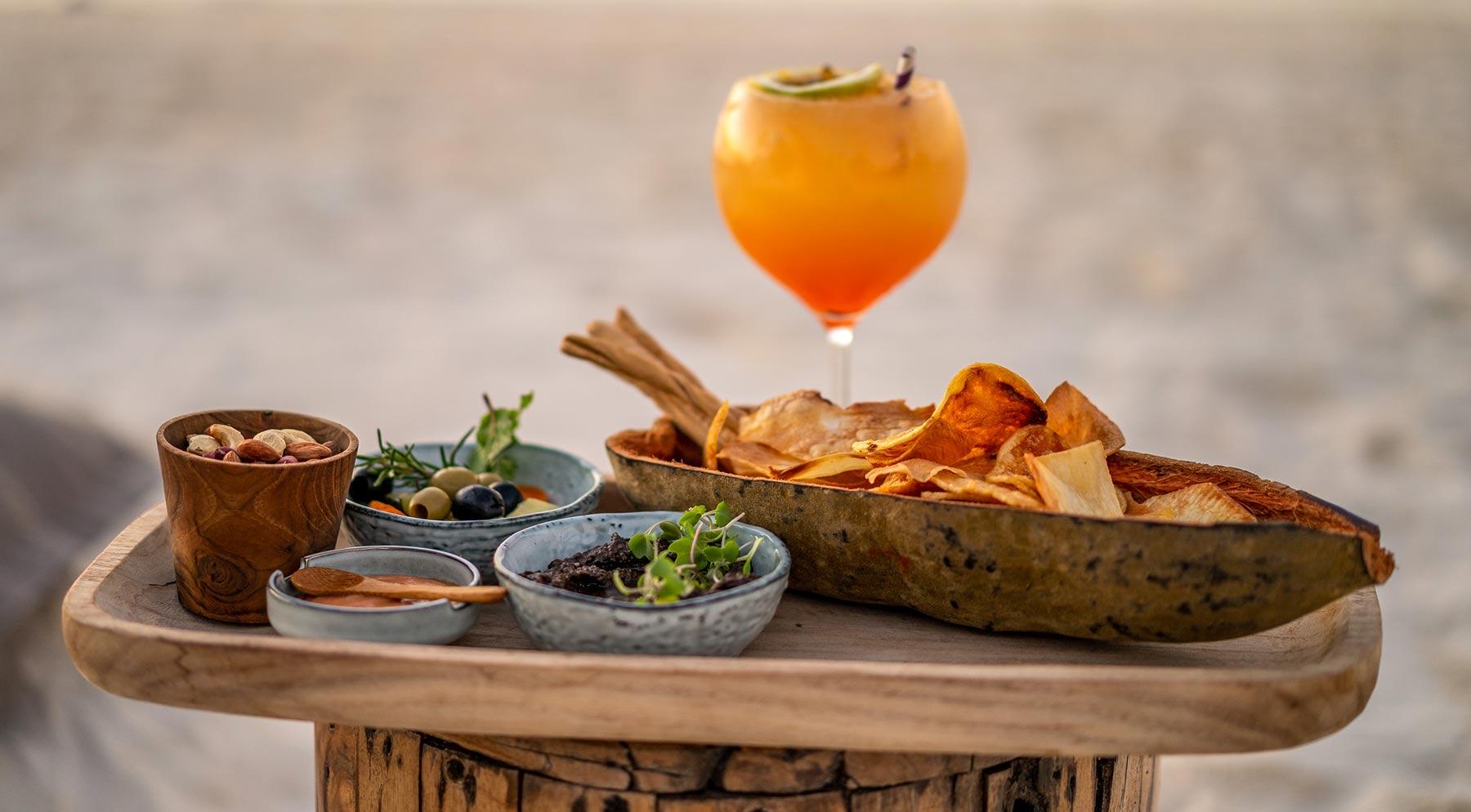 a wooden platter of crisps, olives, nuts and dips beside a glass of an orange cocktail, set on a stand on the beach