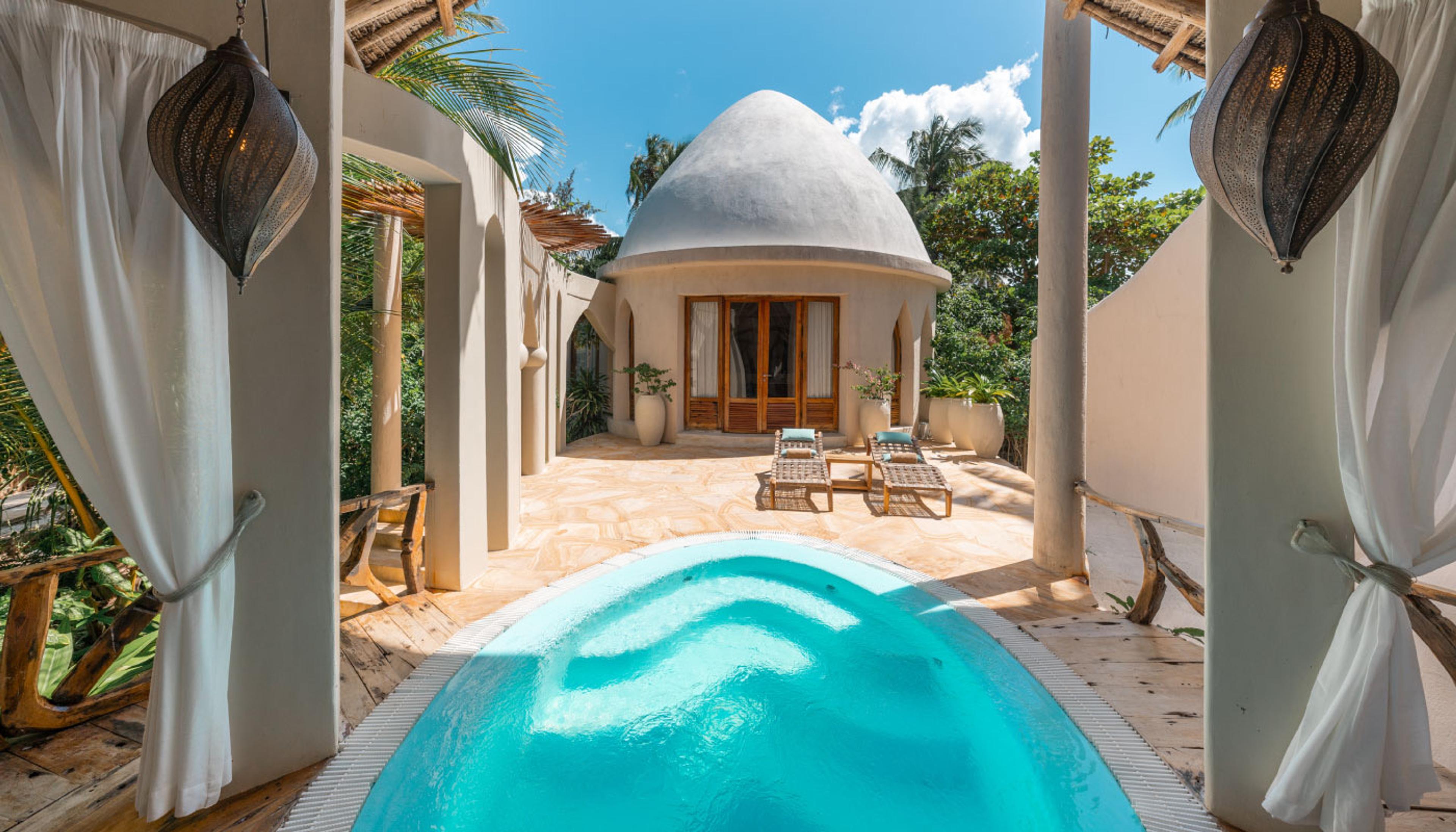 An oval plunge pool on a terrace leading up to a domed, whitewashed villa under a bright sky.