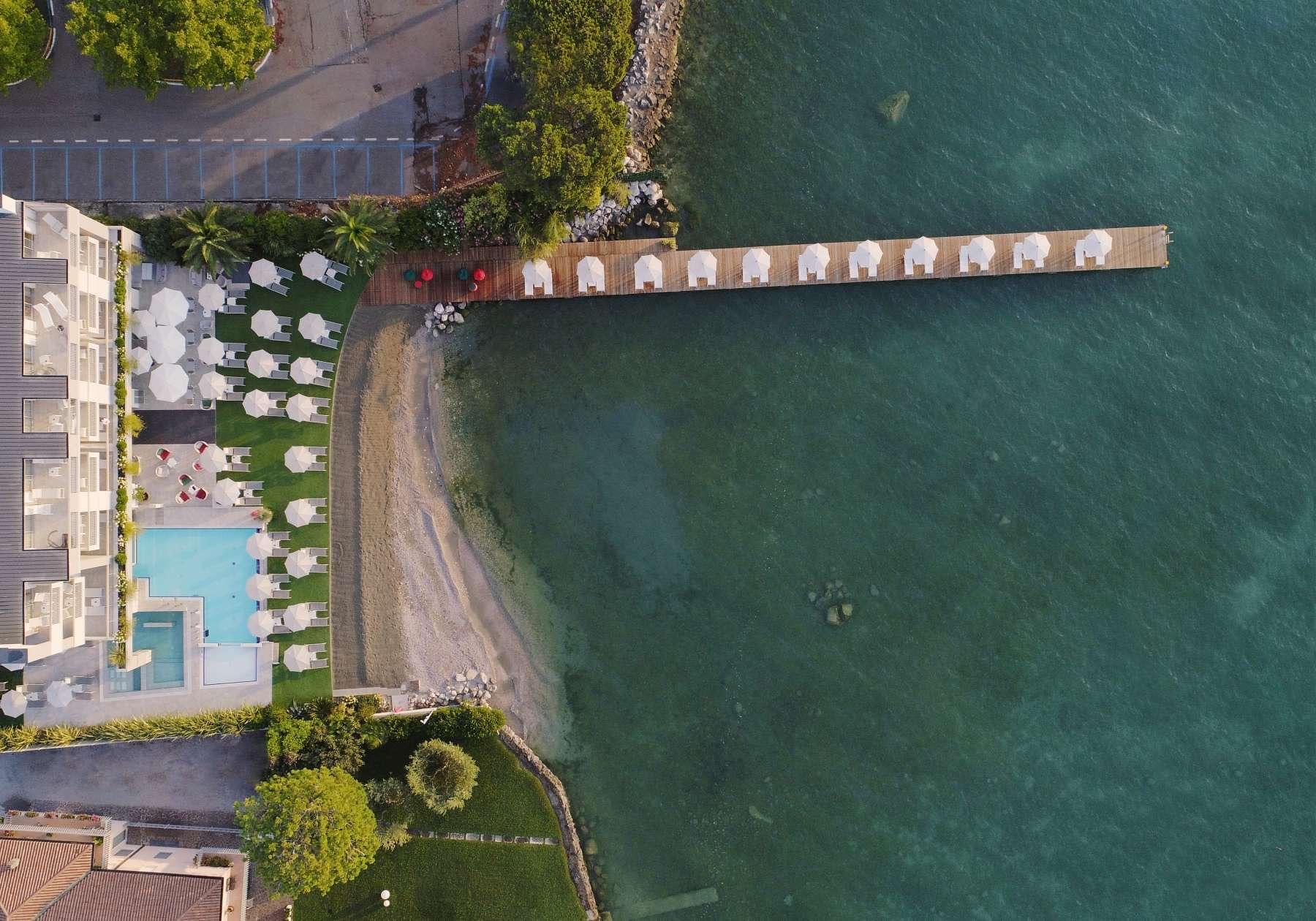 An aerial view of the hotel's lakefront building, pool terrace, beach and dock, with umbrellas lining the shore and the lake stretching away.