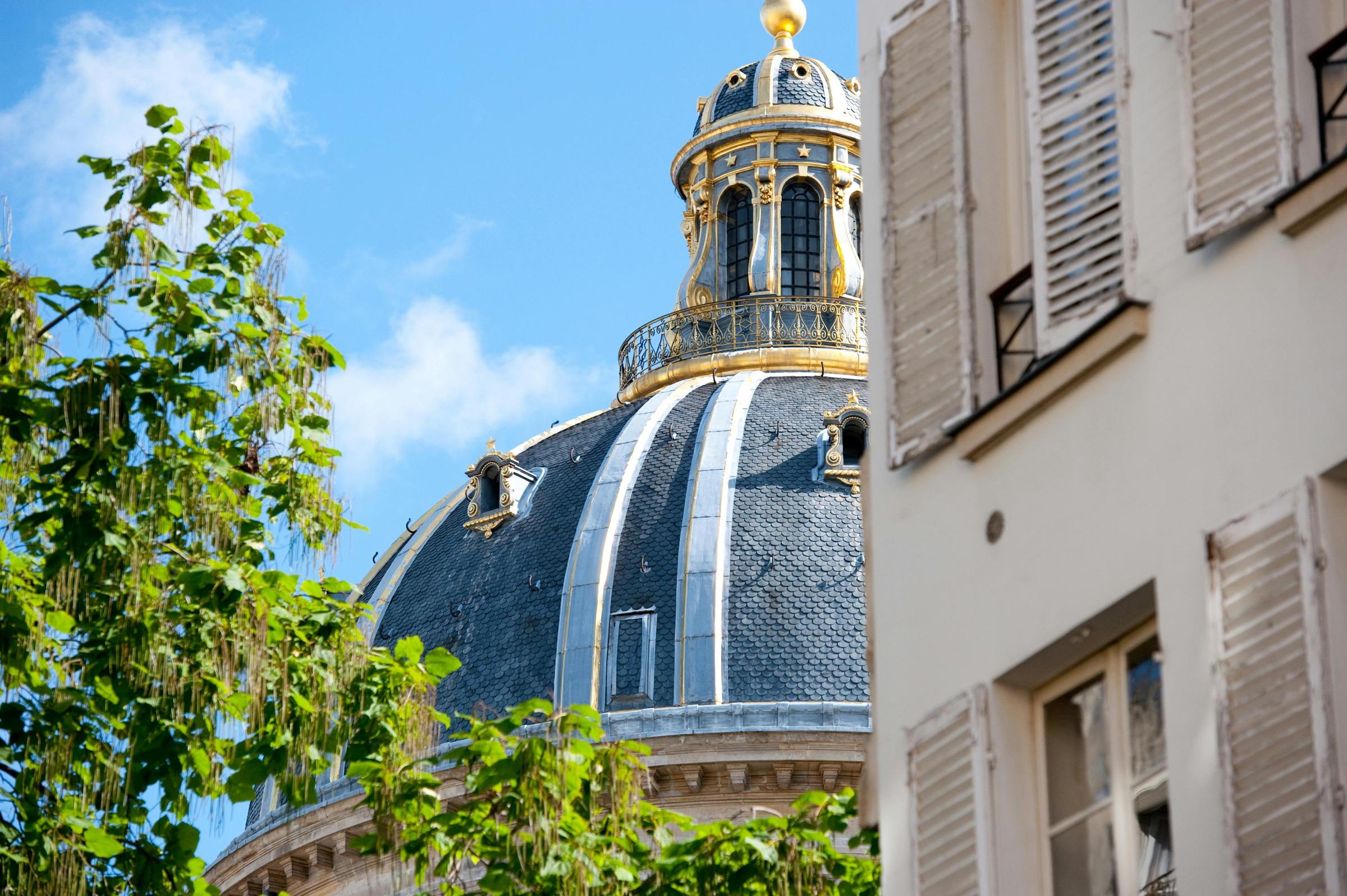 A gilded domed tower rooftop seen past tree branches and a shuttered window, against a blue sky with clouds.
