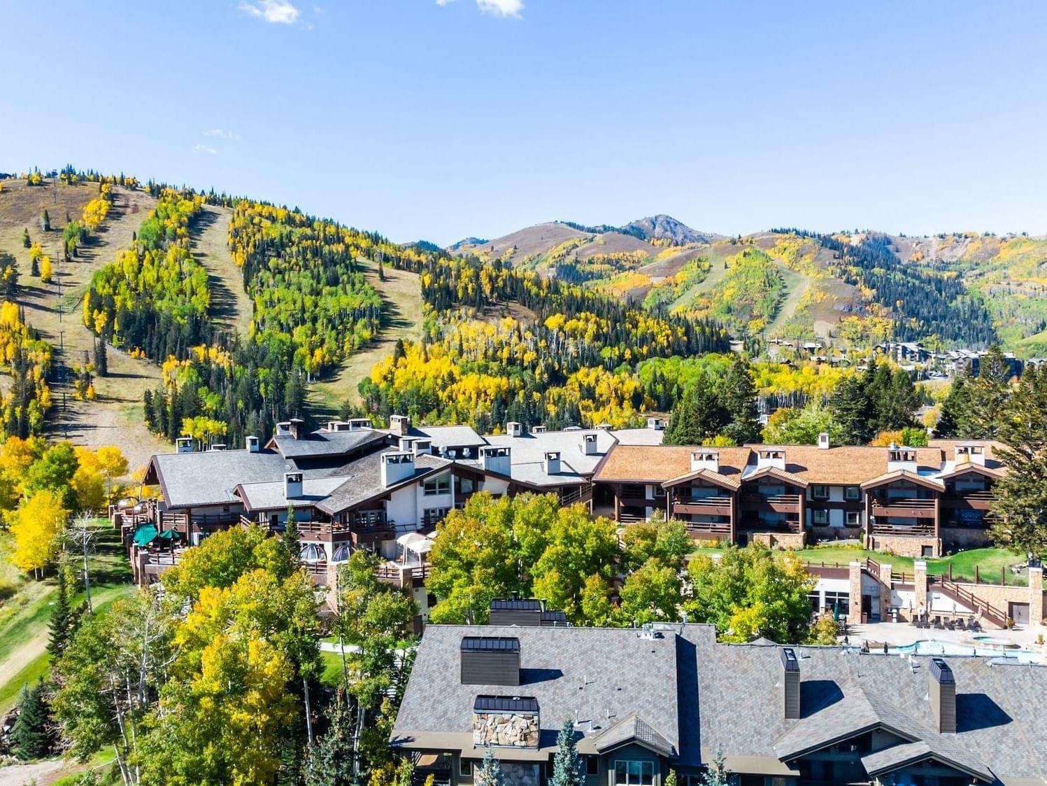 an aerial view of a sprawling timber lodge complex surrounded by golden autumn trees, with ski runs and mountains rising behind it and a pool deck visible at its base