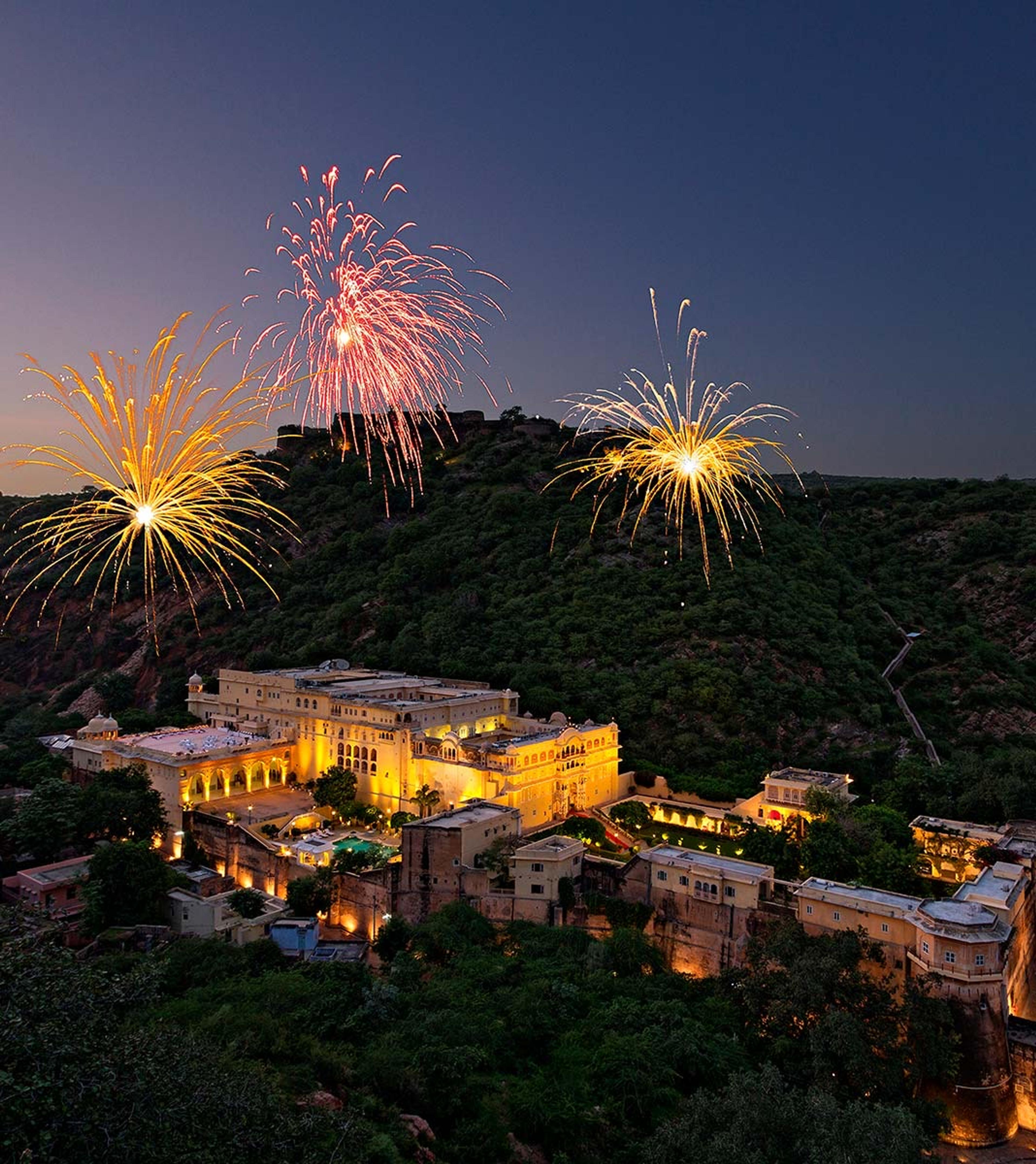 An aerial view at dusk of the palace lit up on a hillside, with fireworks bursting above it.