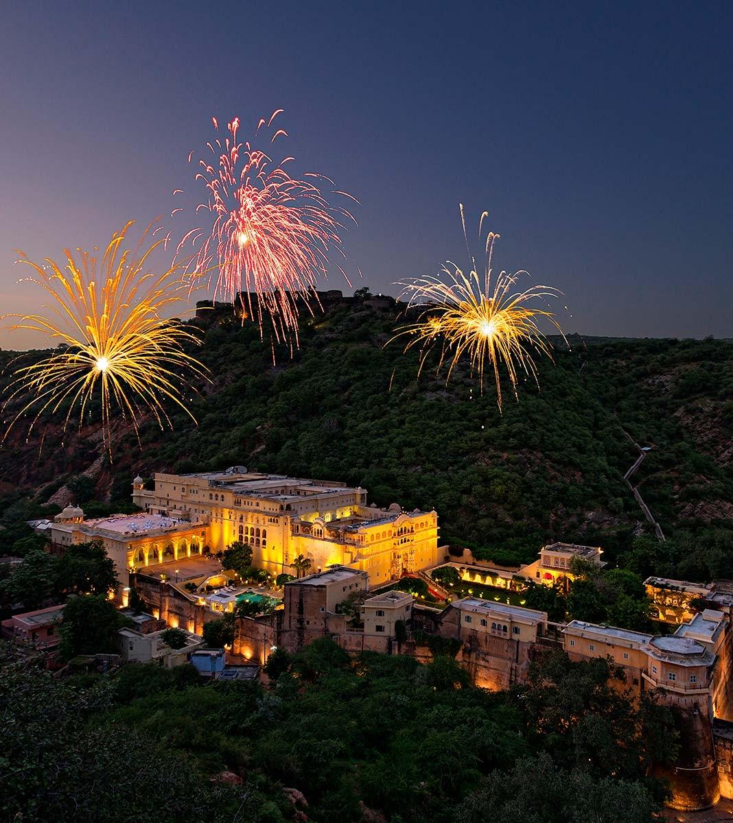 An aerial view at dusk of the palace lit up on a hillside, with fireworks bursting above it.