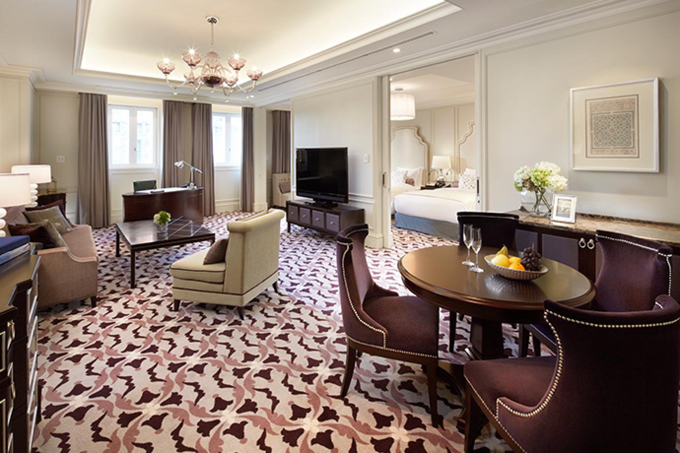 A hotel suite with a round table and chairs set with fruit and wine glasses, a cream sofa and armchair, a writing desk, a wall-mounted television, and an open doorway showing a bed in the adjoining room.