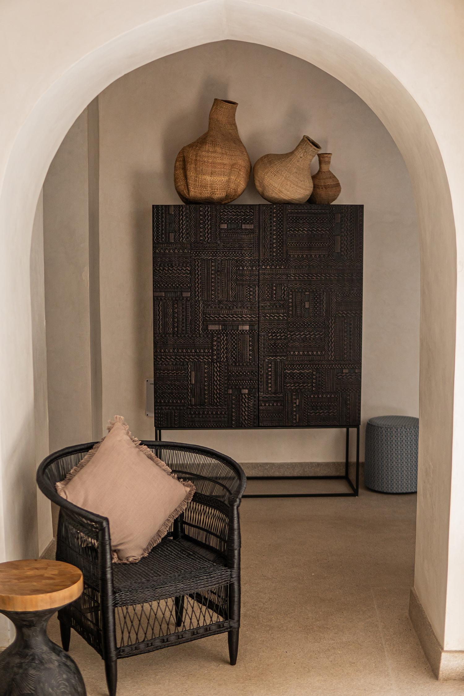 A carved black cabinet topped with three woven vases stands beyond an archway, with a black rattan armchair in the foreground.
