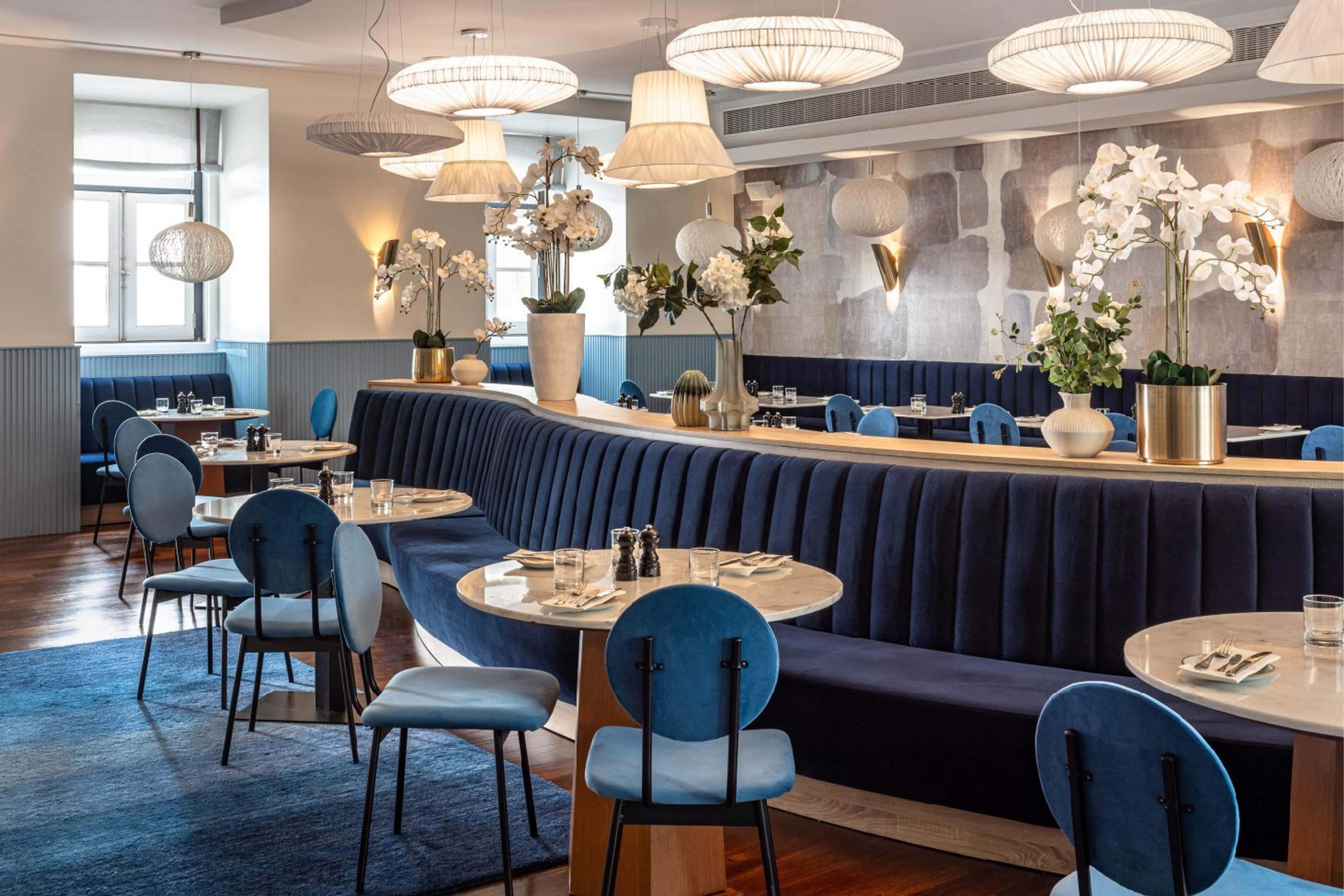 A restaurant with a curved navy velvet banquette, round marble-topped tables, pendant lamps and white orchids.